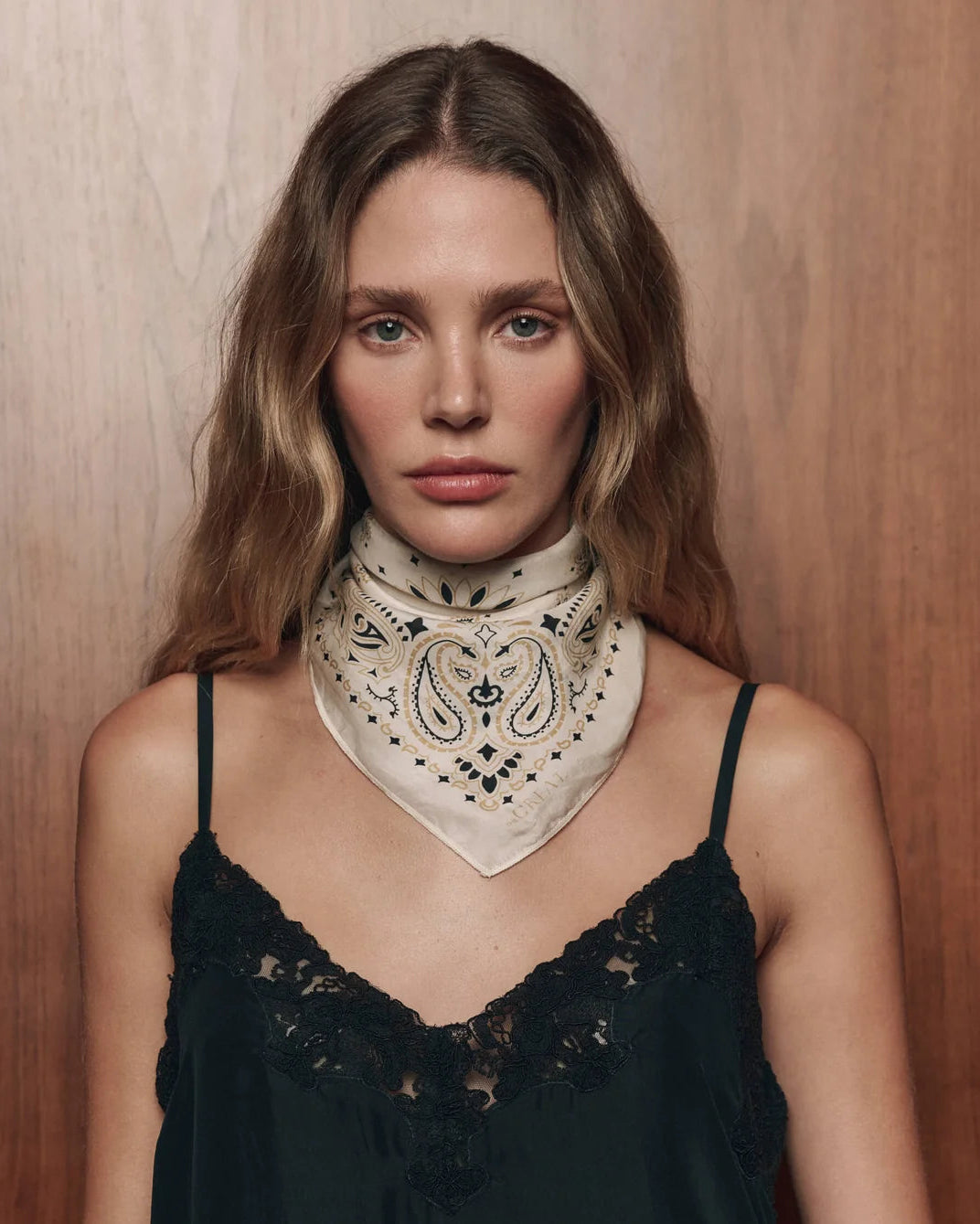 A woman with wavy light brown hair wears a cream-colored paisley bandana tied around her neck over a black lace camisole top, set against a warm wood-paneled background.