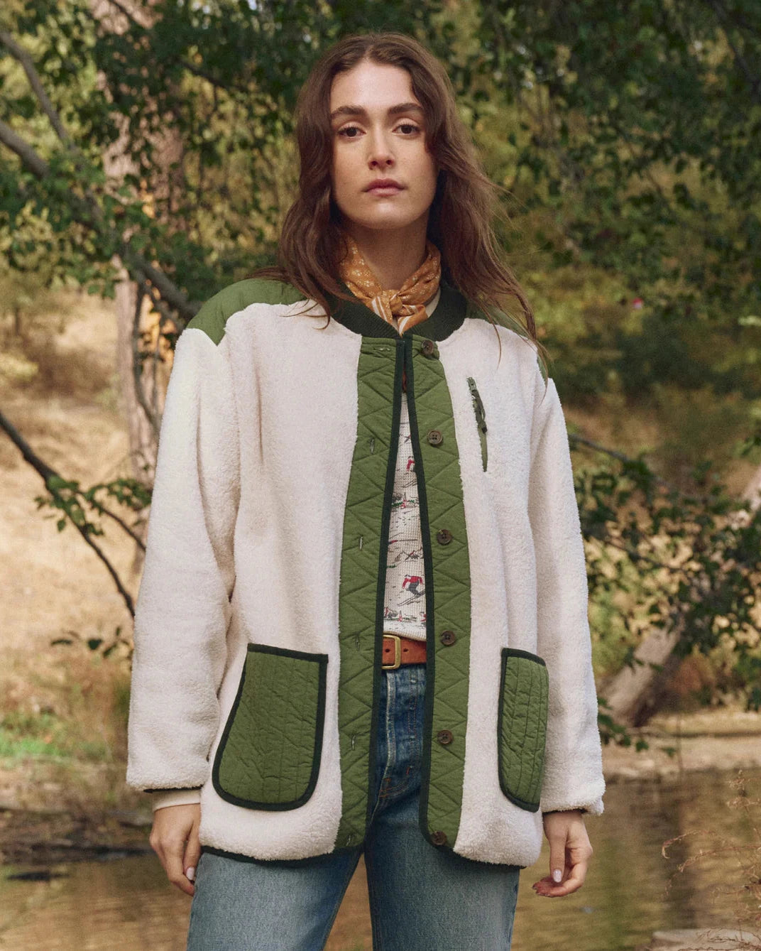 Woman wearing a white and green jacket in a natural setting with trees and water.
