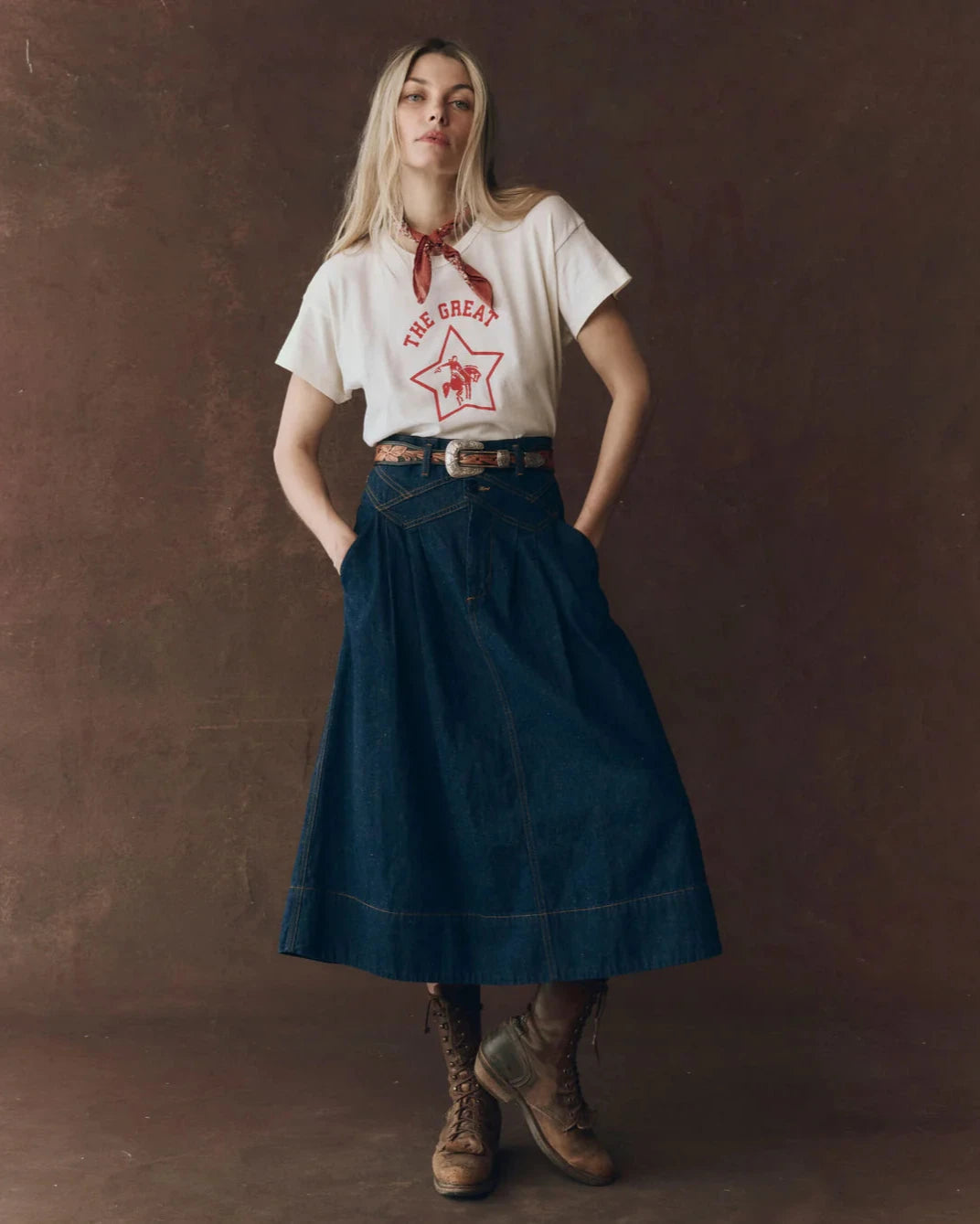 Woman wearing a white t-shirt with text and graphics, a blue skirt, and brown boots against a dark background.