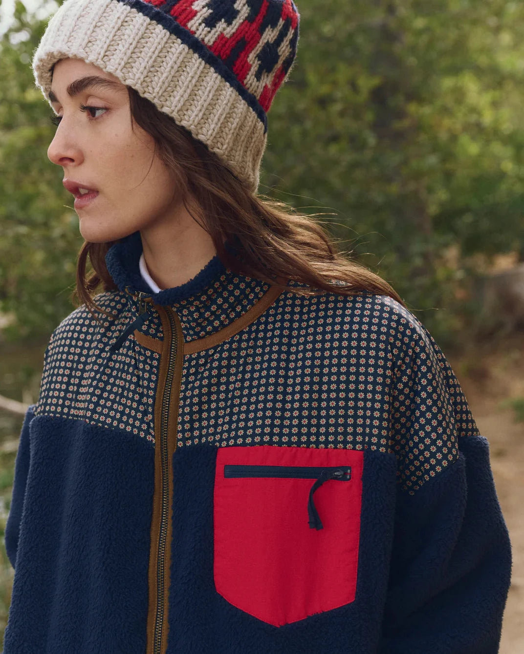 Person wearing a patterned jacket with a red pocket and a knit hat outdoors.