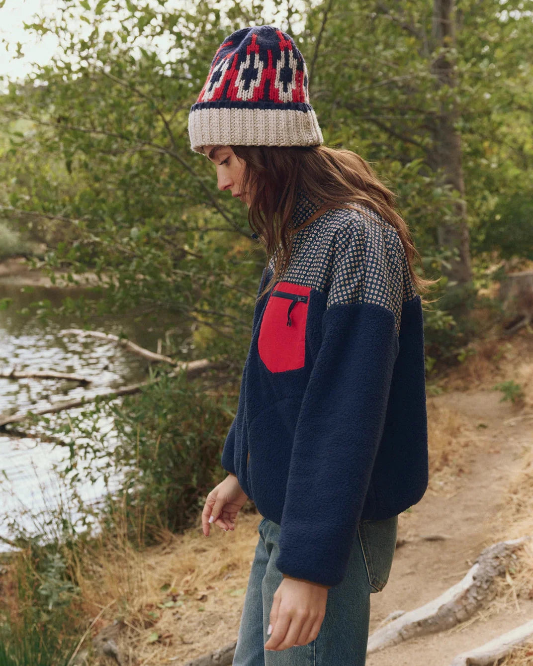 Person wearing a patterned beanie and a navy sweater with a red logo, standing outdoors by a body of water.