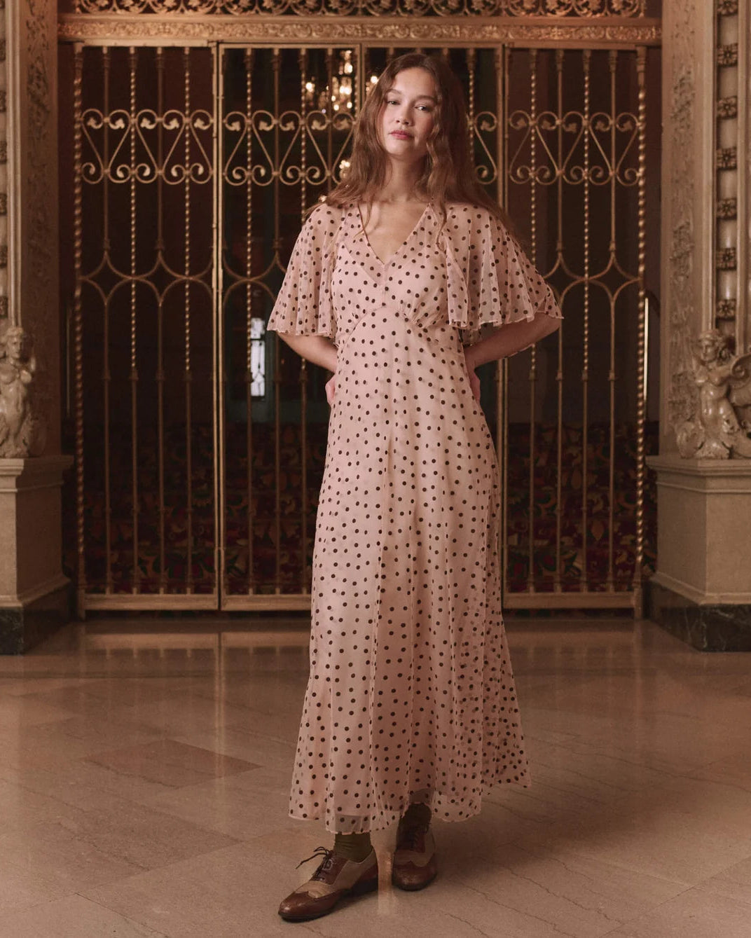 model wearing pink dress semi sheer dress with polka dots behind a ornate metal background.
