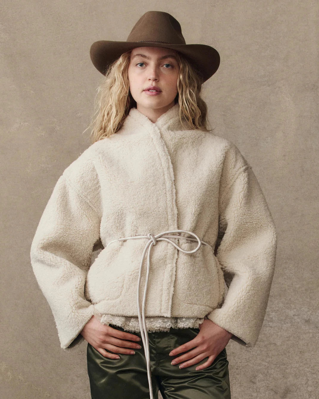 Person wearing a cream sherpa jacket and brown hat against a beige background