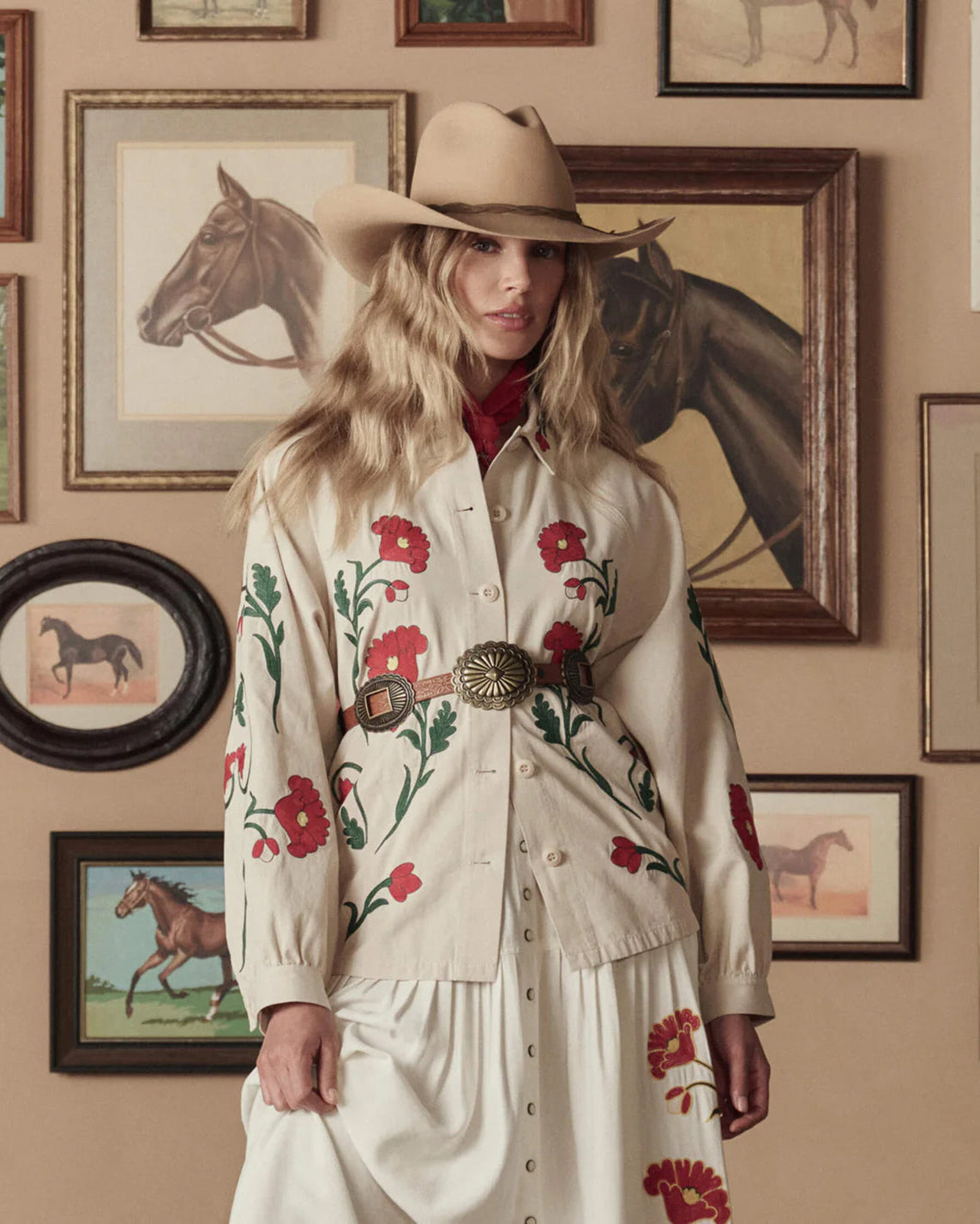 Woman wearing a floral embroidered shirt and cowboy hat in front of framed horse pictures.