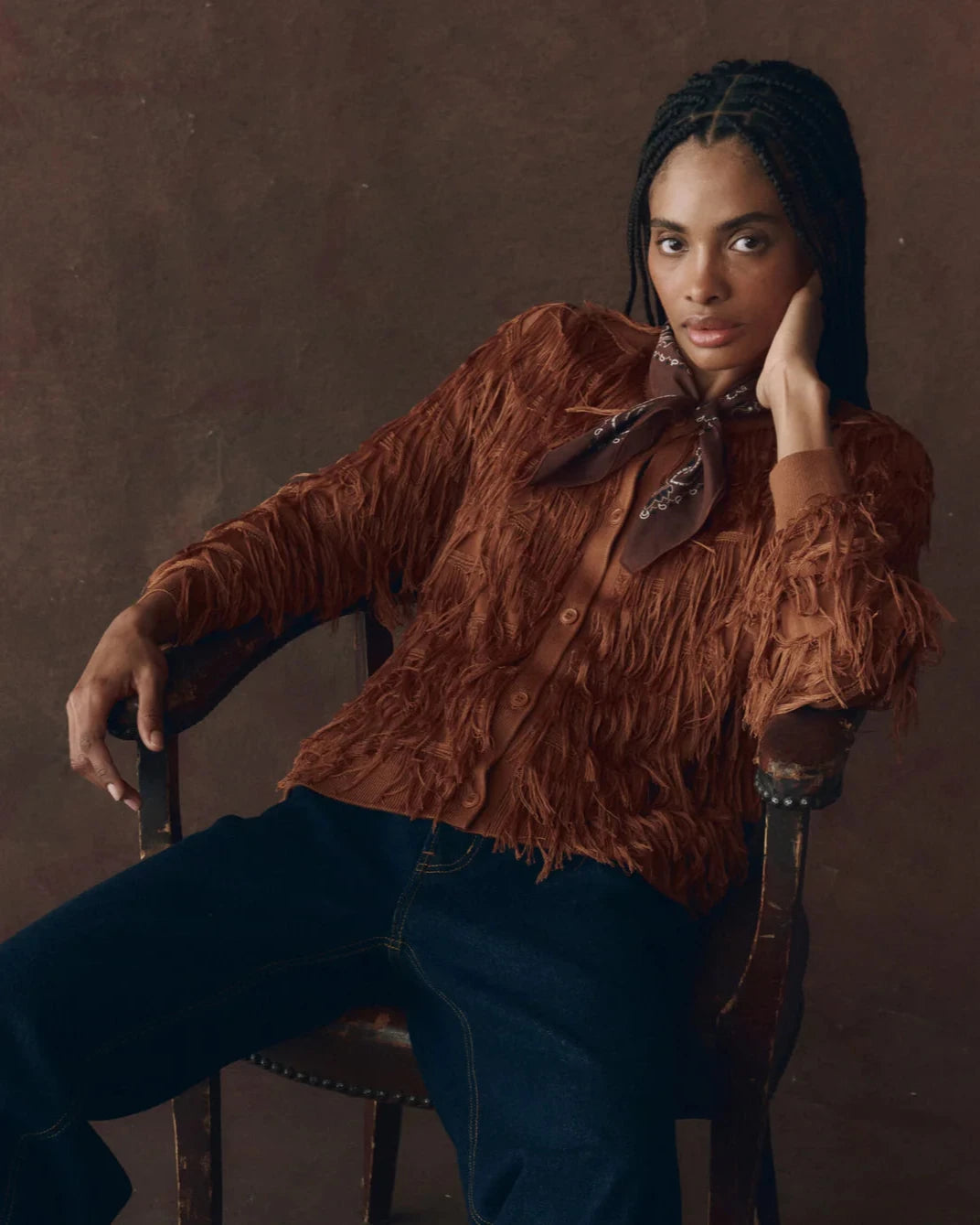 Person wearing a brown textured shirt with a bow tie, sitting on a chair against a brown background