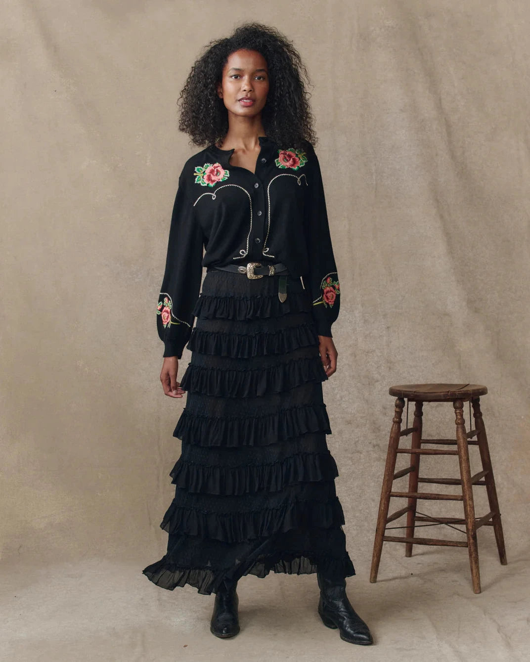 Woman wearing a black embroidered shirt and ruffled skirt standing next to a wooden stool against a beige background.