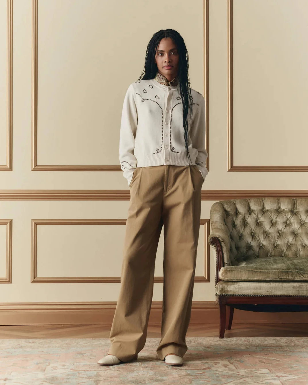 Woman in a stylish outfit standing in an elegant room with a tufted sofa.