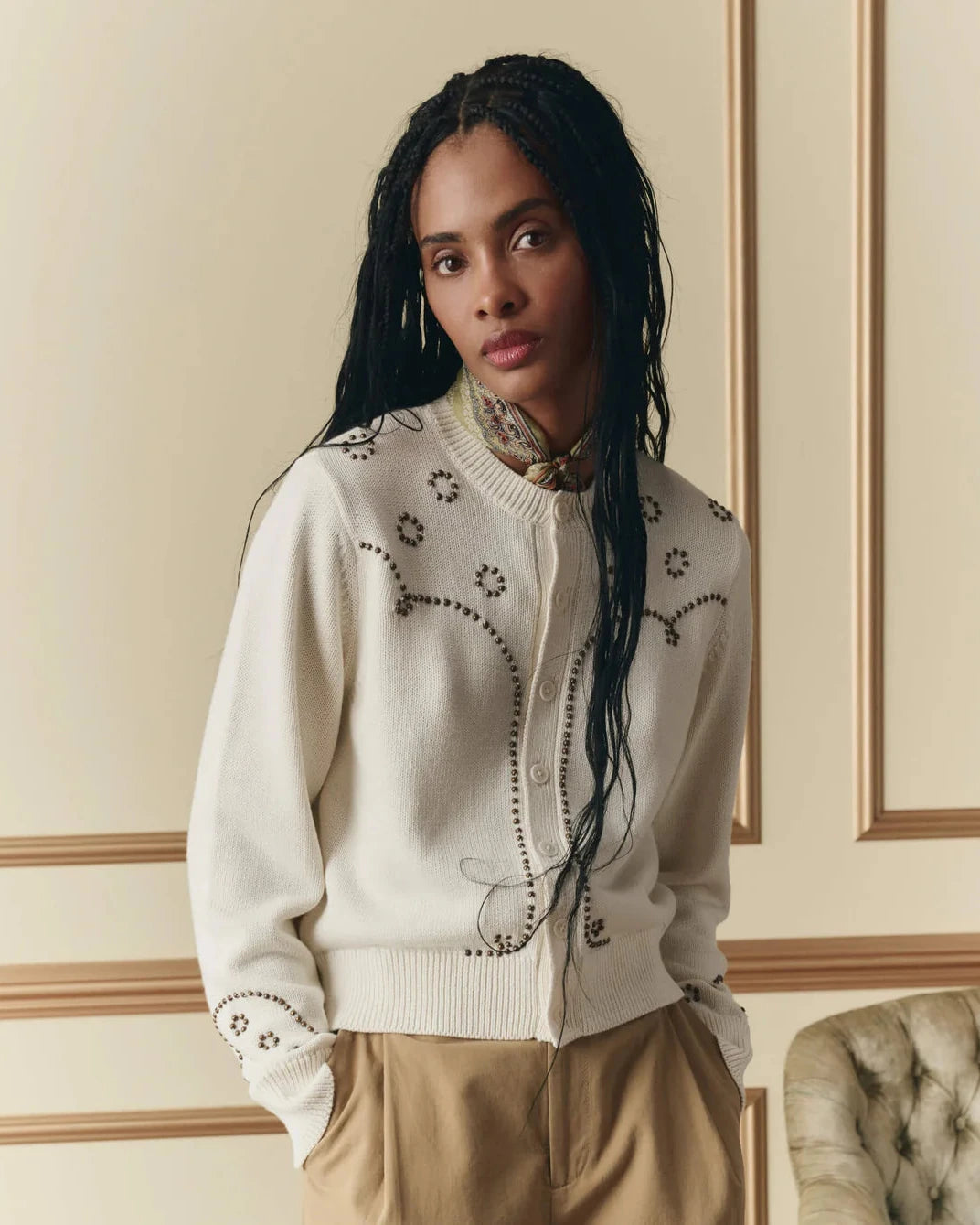 Woman wearing a cream-colored cardigan with floral patterns in a room with decorative wall molding.
