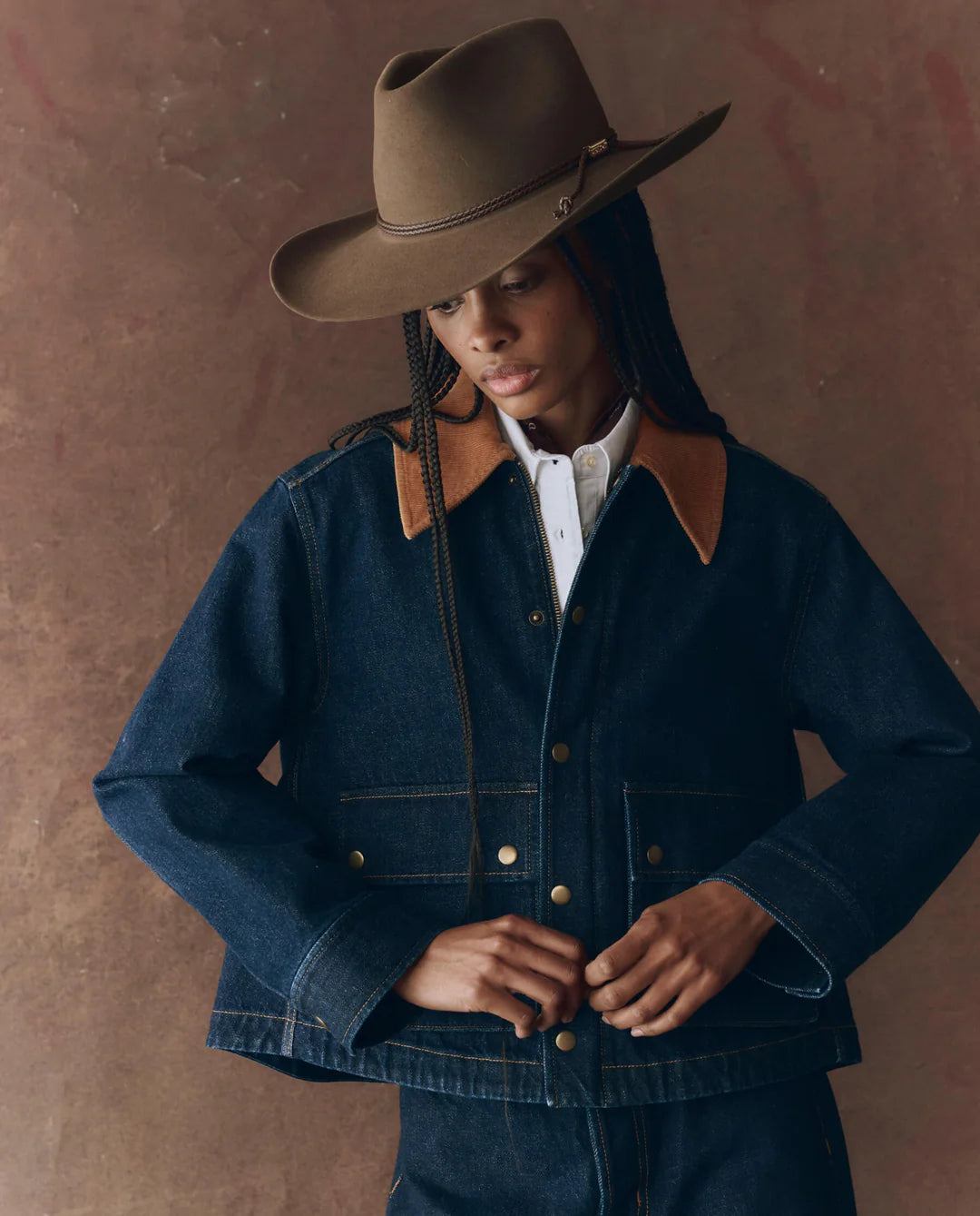 Person wearing a dark blue denim jacket with brown leather accents and a brown cowboy hat against a brown background.