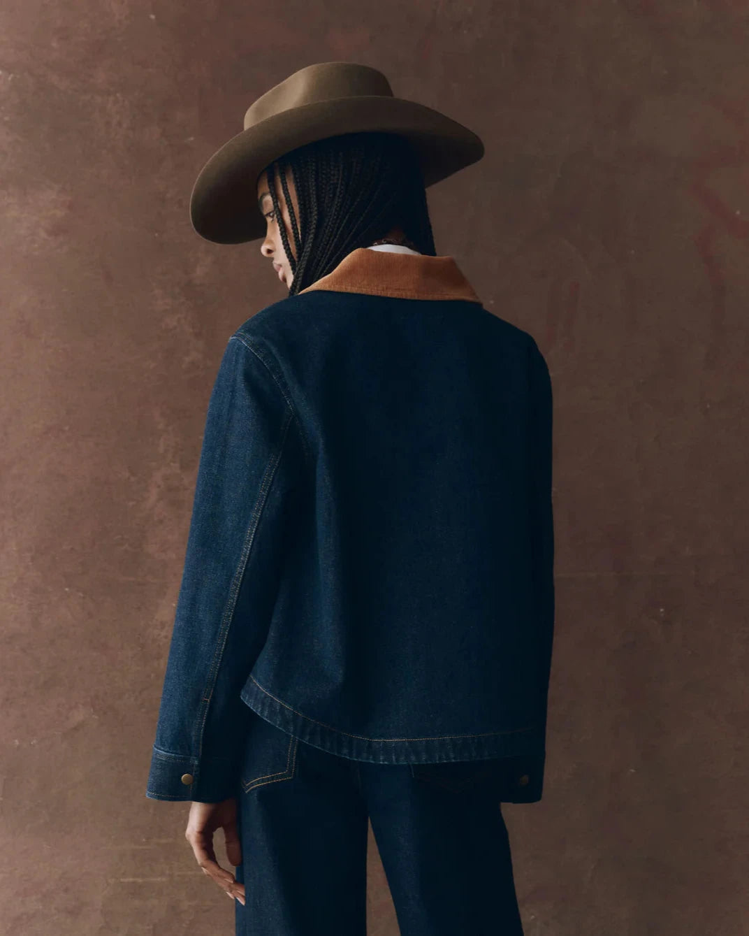 Person wearing a denim jacket and wide-brimmed hat against a brown background
