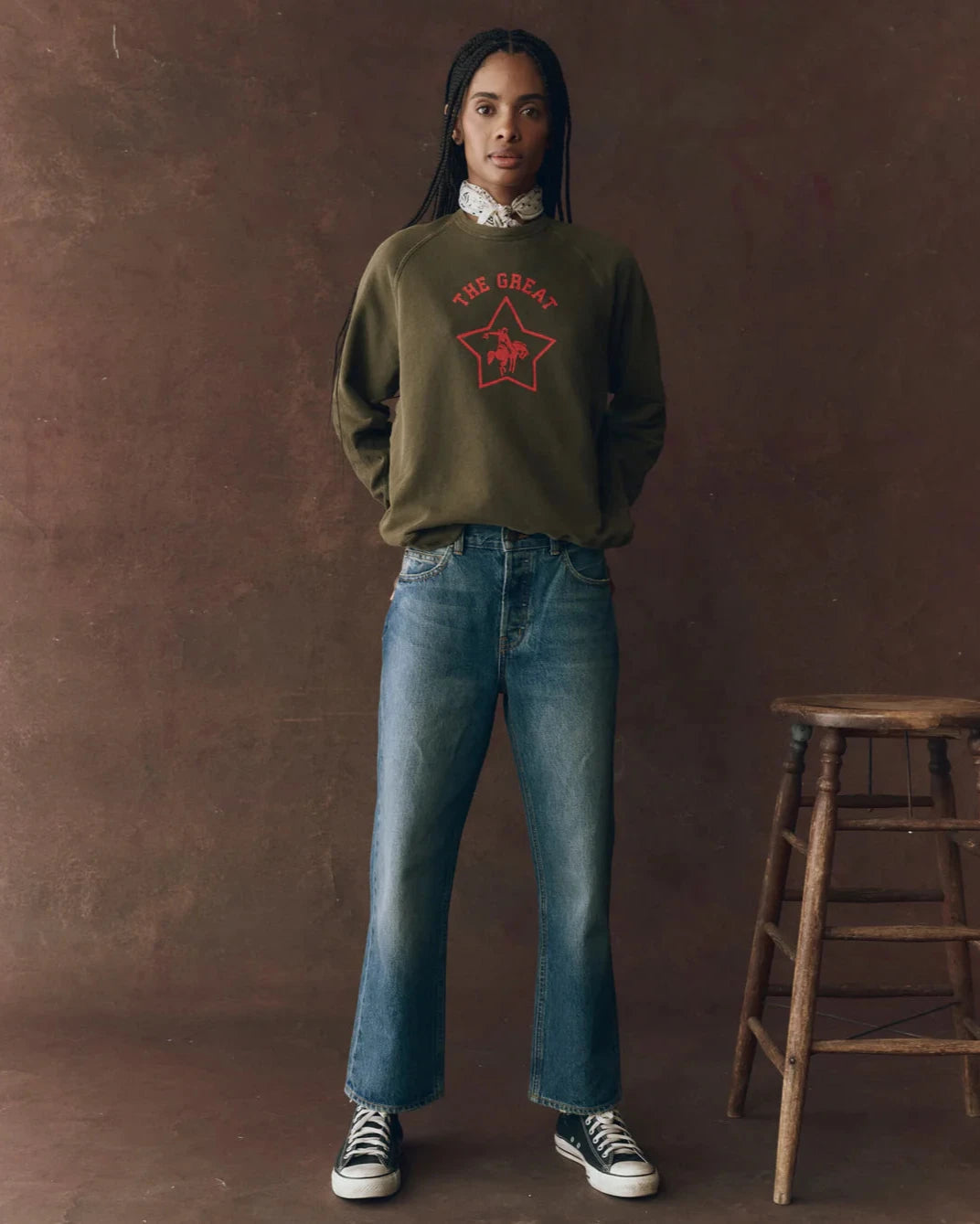 Person wearing a green sweatshirt with a red logo and blue jeans against a brown background