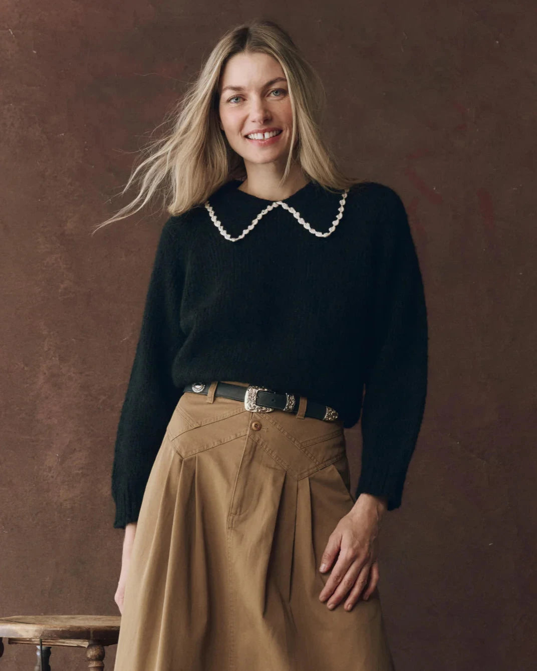 Woman wearing a black sweater with a white collar and beige pants against a brown background