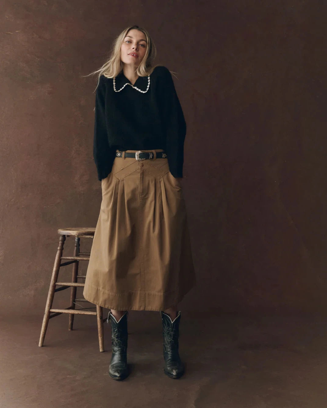Woman in a black sweater and beige skirt standing against a brown background