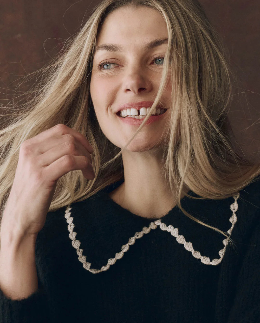 Woman with blonde hair wearing a black sweater with a white collar against a brown background
