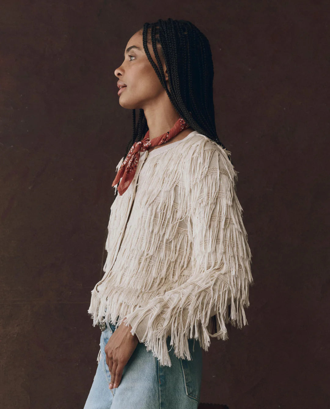 Woman wearing a white fringed jacket and blue jeans against a brown background