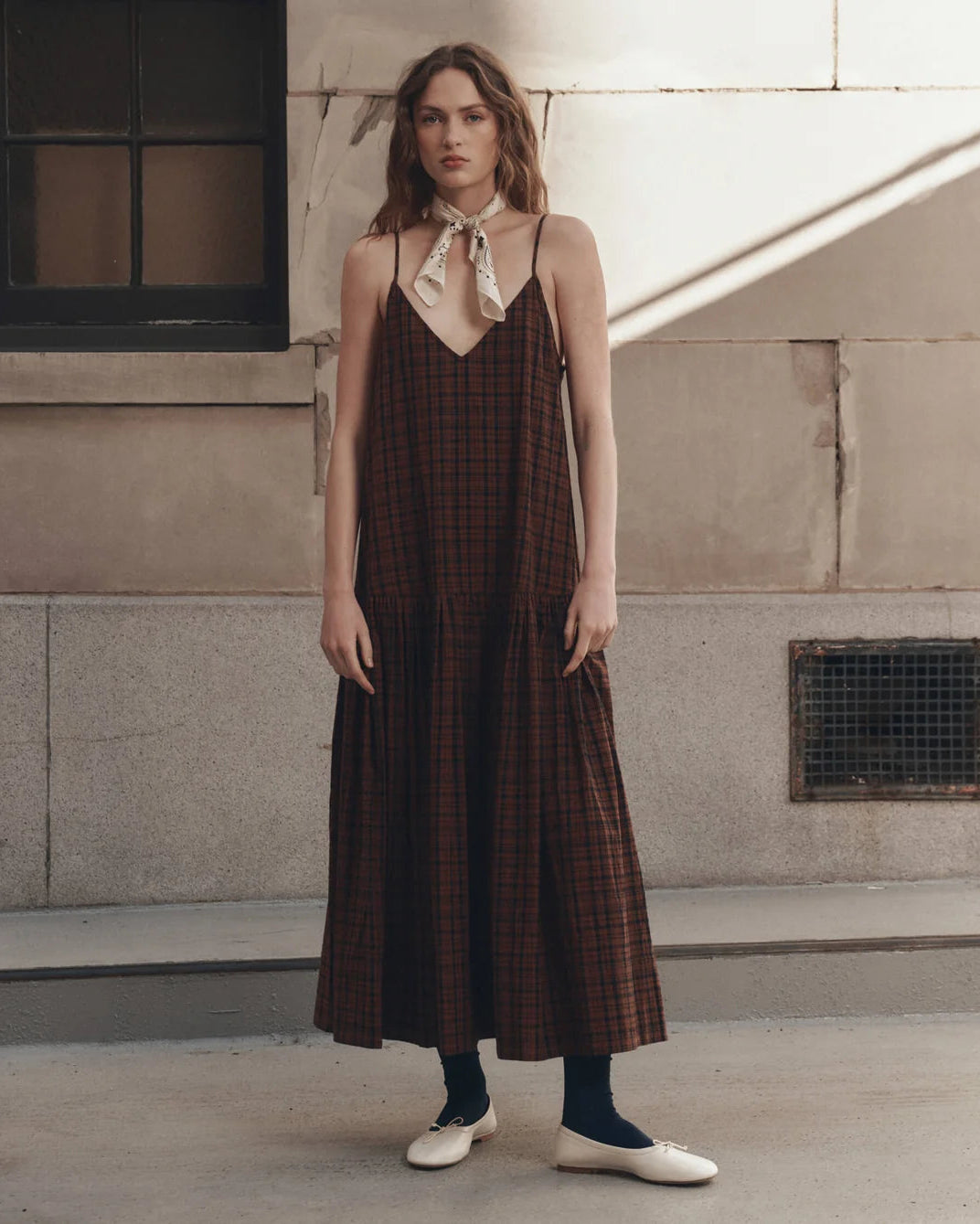 A woman stands outdoors against a stone building wall wearing a dark brown plaid midi slip dress with thin straps and a cream neck scarf, paired with black socks and white shoes.