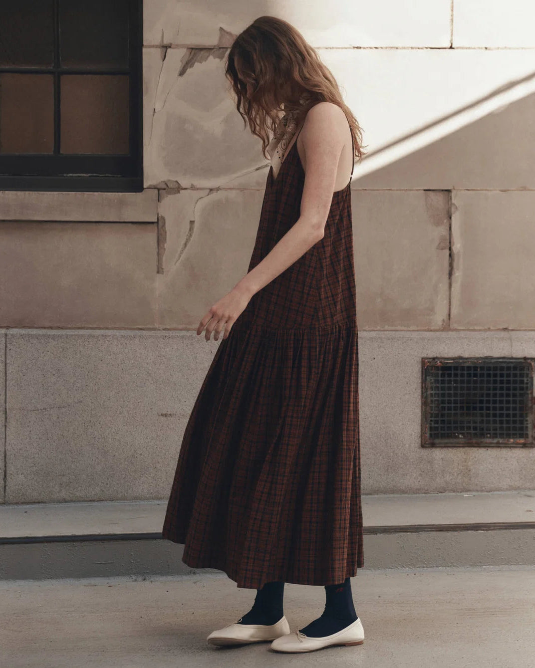 A woman wearing a brown and red plaid sleeveless midi dress, black socks, and white flat shoes, standing outdoors against a textured concrete wall.