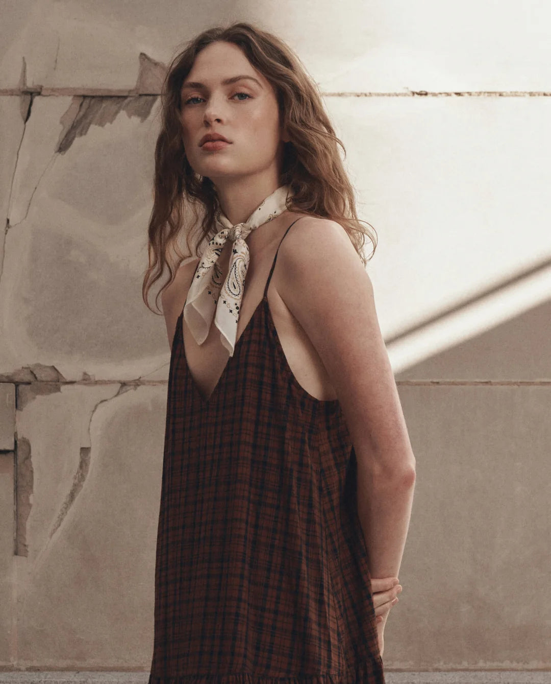 A woman wearing a dark brown and black plaid halter dress with a cream paisley neck scarf, standing against a textured beige plaster wall.