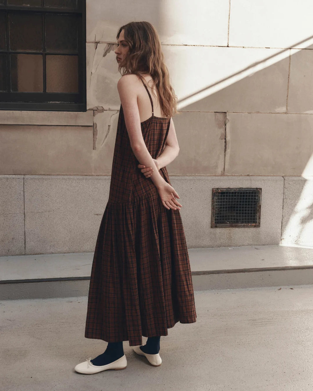 A woman wearing a brown plaid maxi dress with thin straps, standing outdoors against a stone wall.
