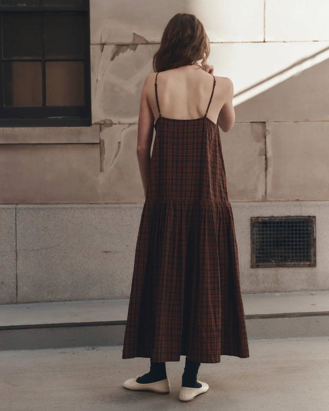 A woman viewed from behind wearing a brown and black plaid midi slip dress with thin spaghetti straps, standing outdoors against a textured concrete wall.