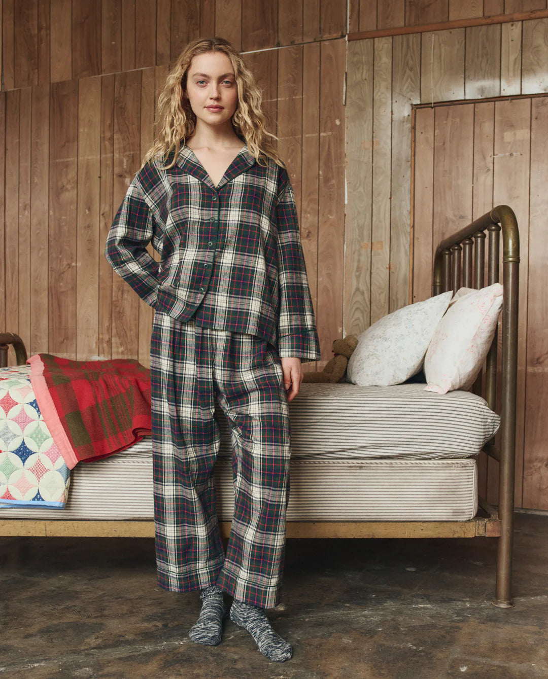 Woman wearing a plaid pajama set in a rustic room with wooden walls and a bed.