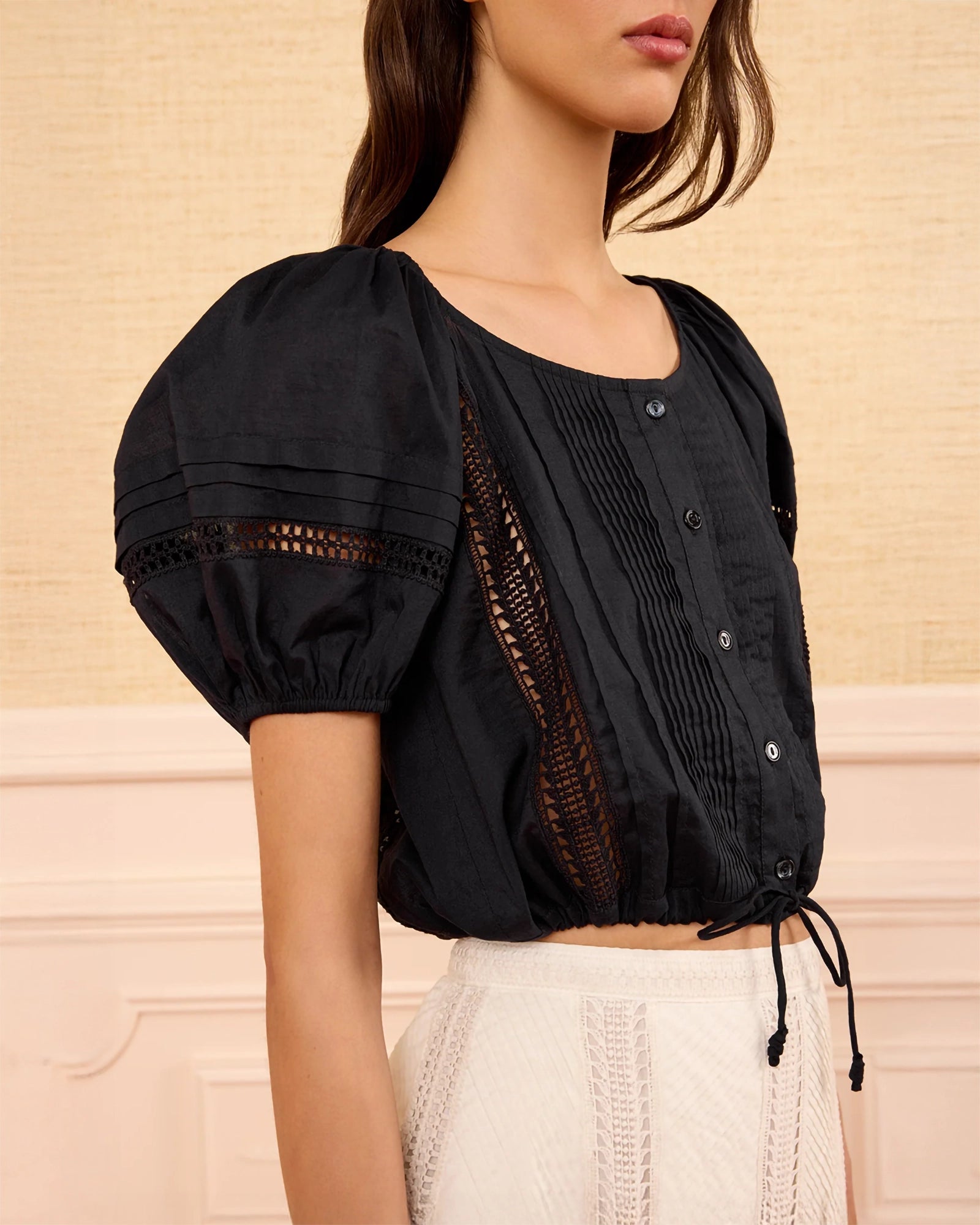 A woman wearing a black short-sleeve blouse with puff sleeves, vertical pleats, and sheer mesh side panels, paired with a white textured skirt against a beige wall.