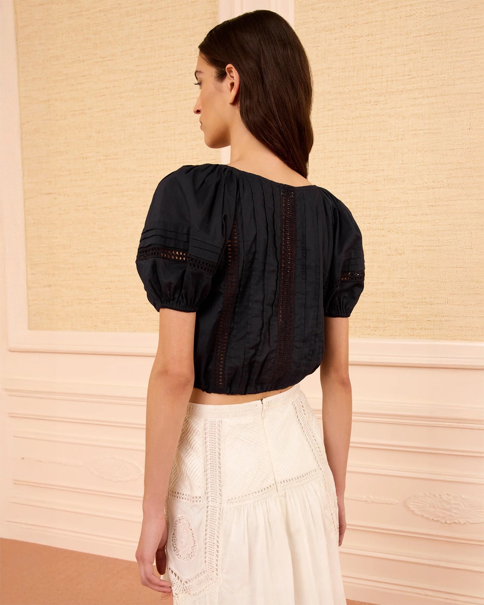 A woman shown from the back wearing a black short-sleeve cropped blouse with puff sleeves and vertical lace-up detailing, paired with a white skirt, set against a cream-colored paneled wall.