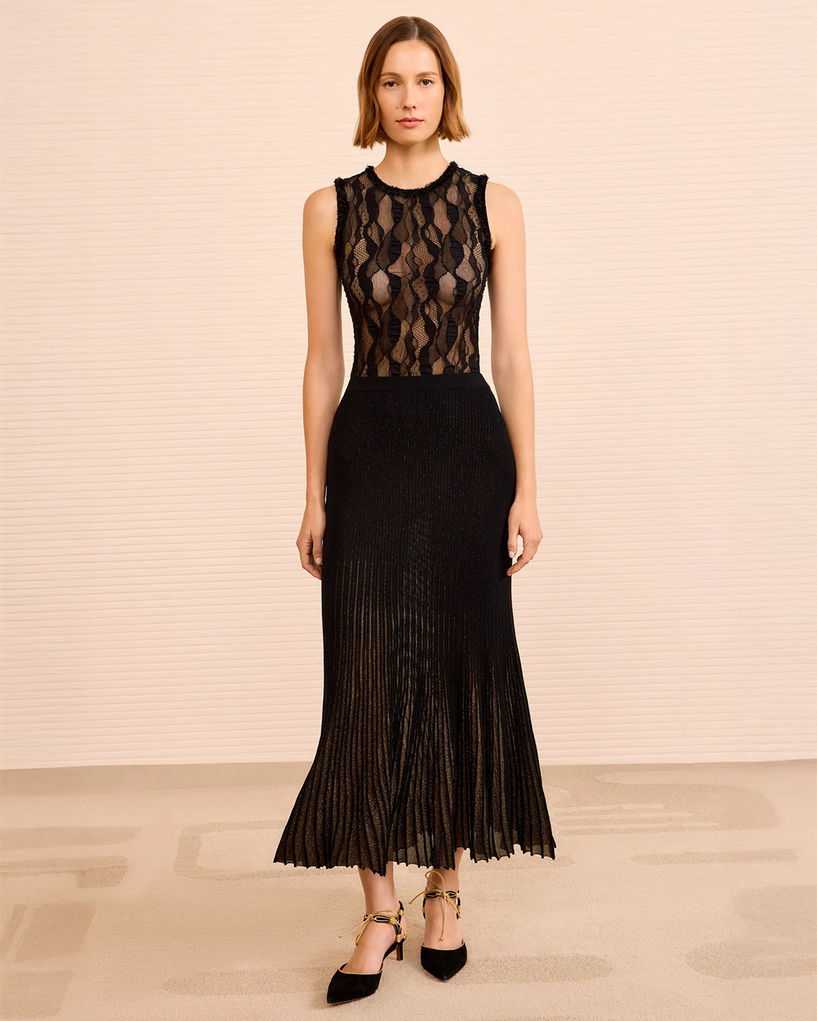 Woman wearing a black lace top with a pleated metallic skirt on the bottom with a beige background
