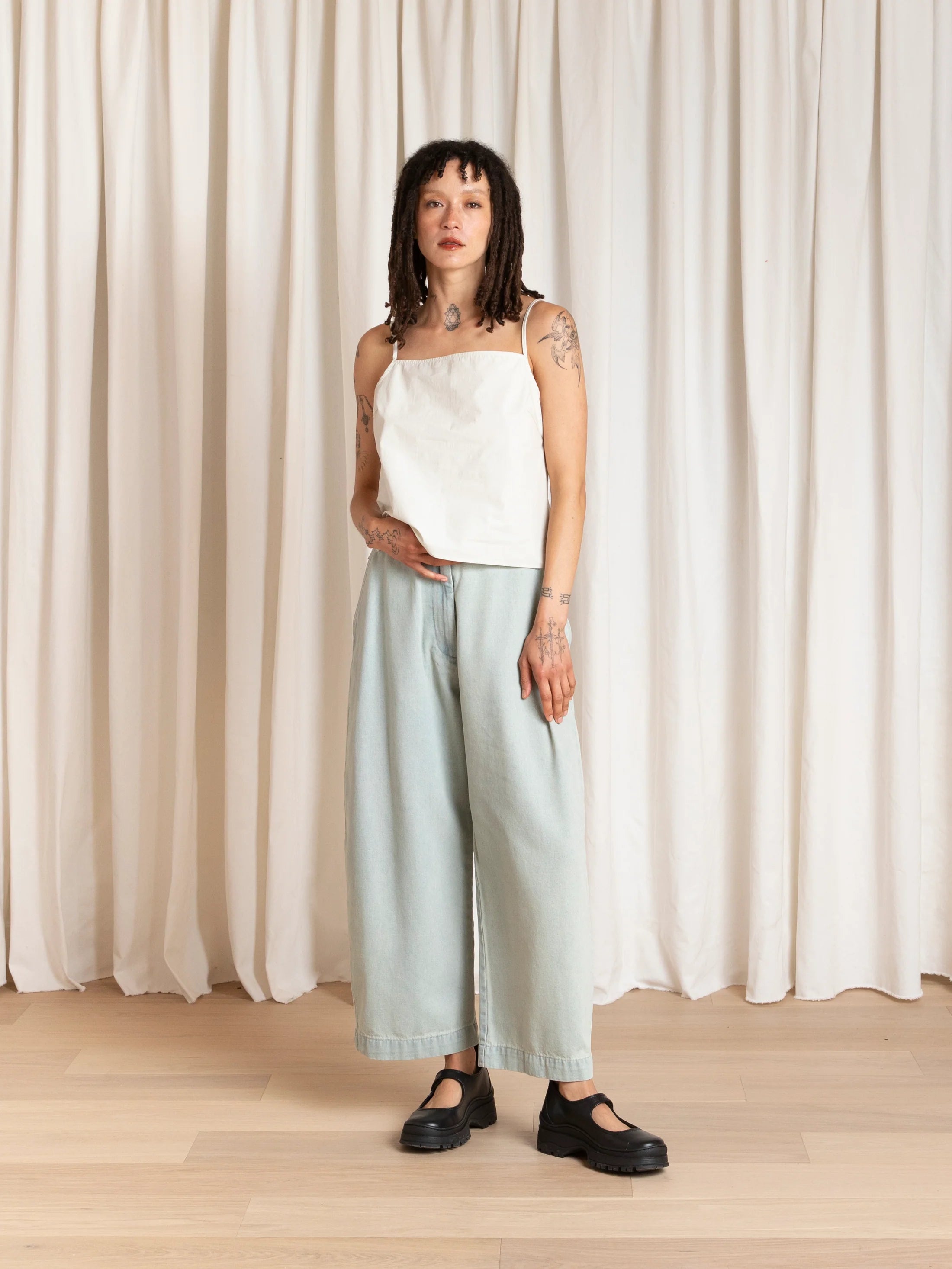 Woman wearing a white top and light blue pants standing in front of white curtains.
