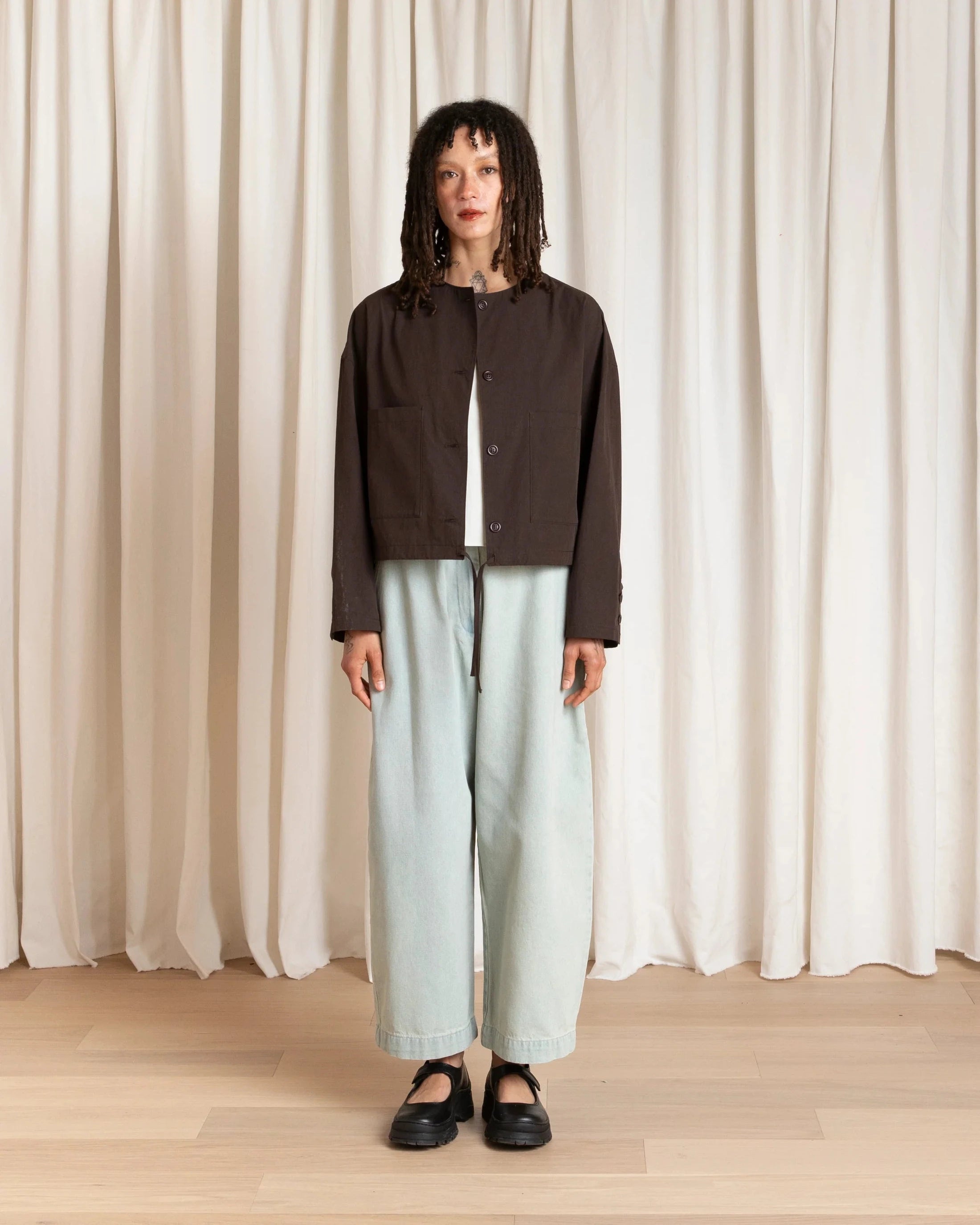 Person wearing a dark jacket and light pants standing against a white curtain.