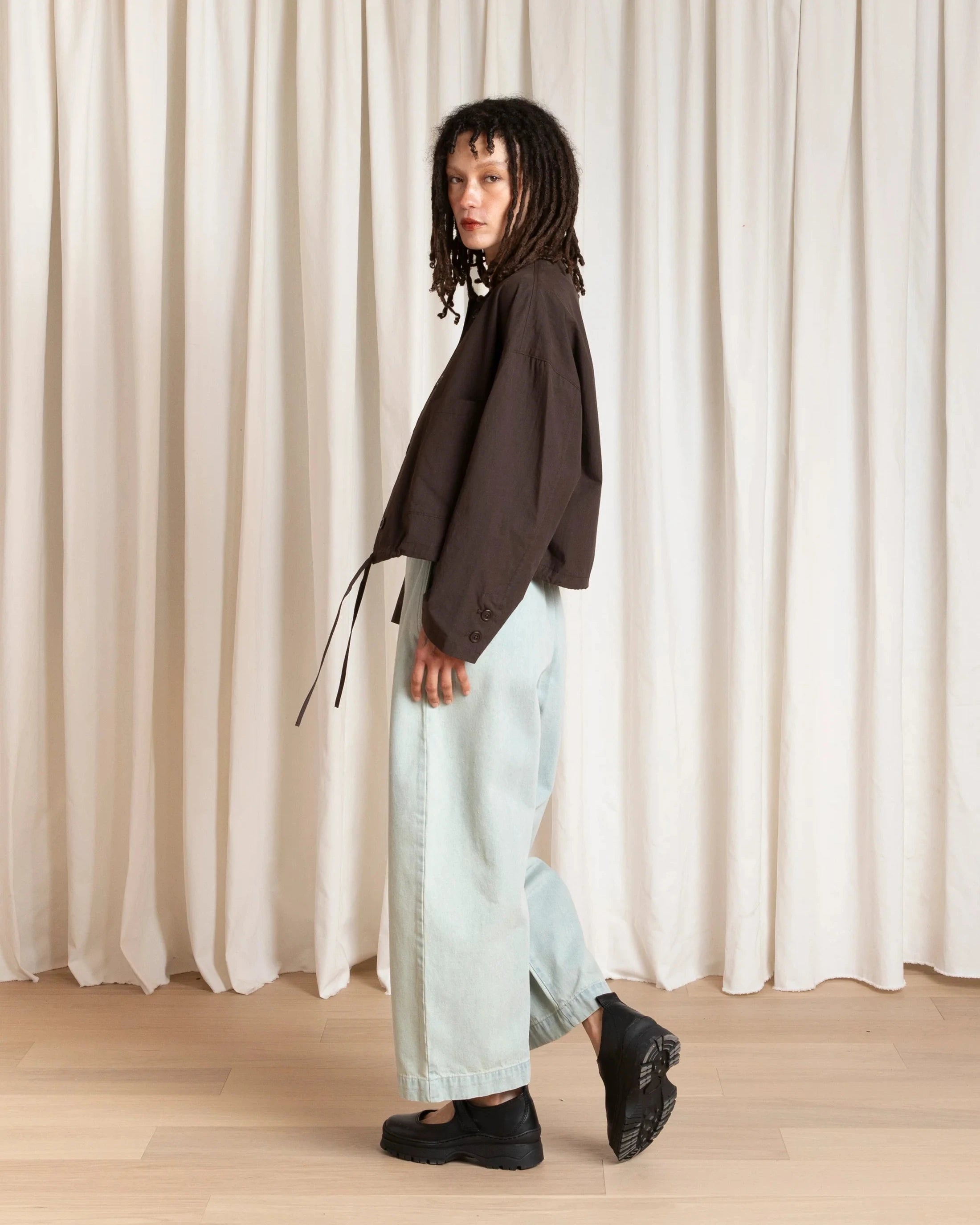 Person wearing a brown top and light blue jeans standing against a white curtain.