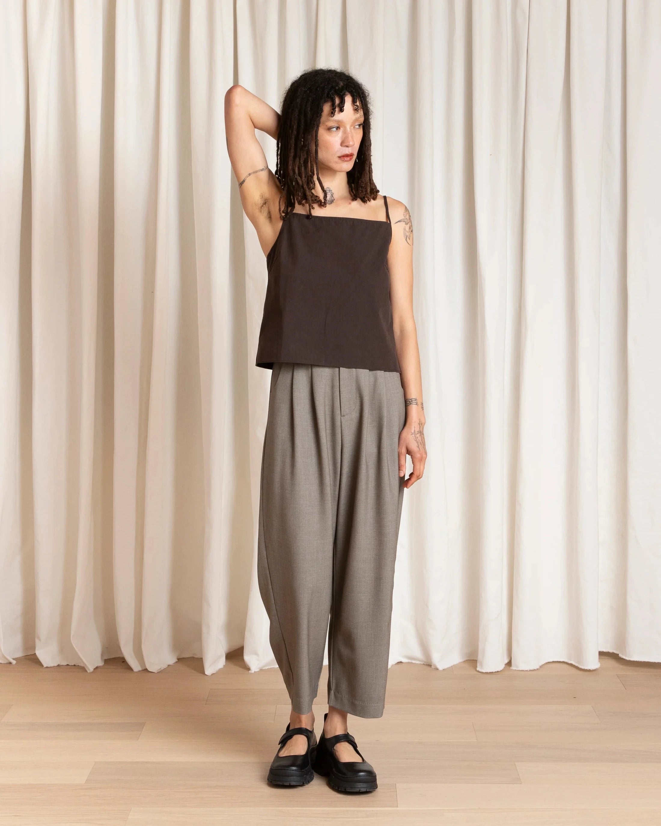 Woman wearing a black top and gray pants standing against a white curtain.