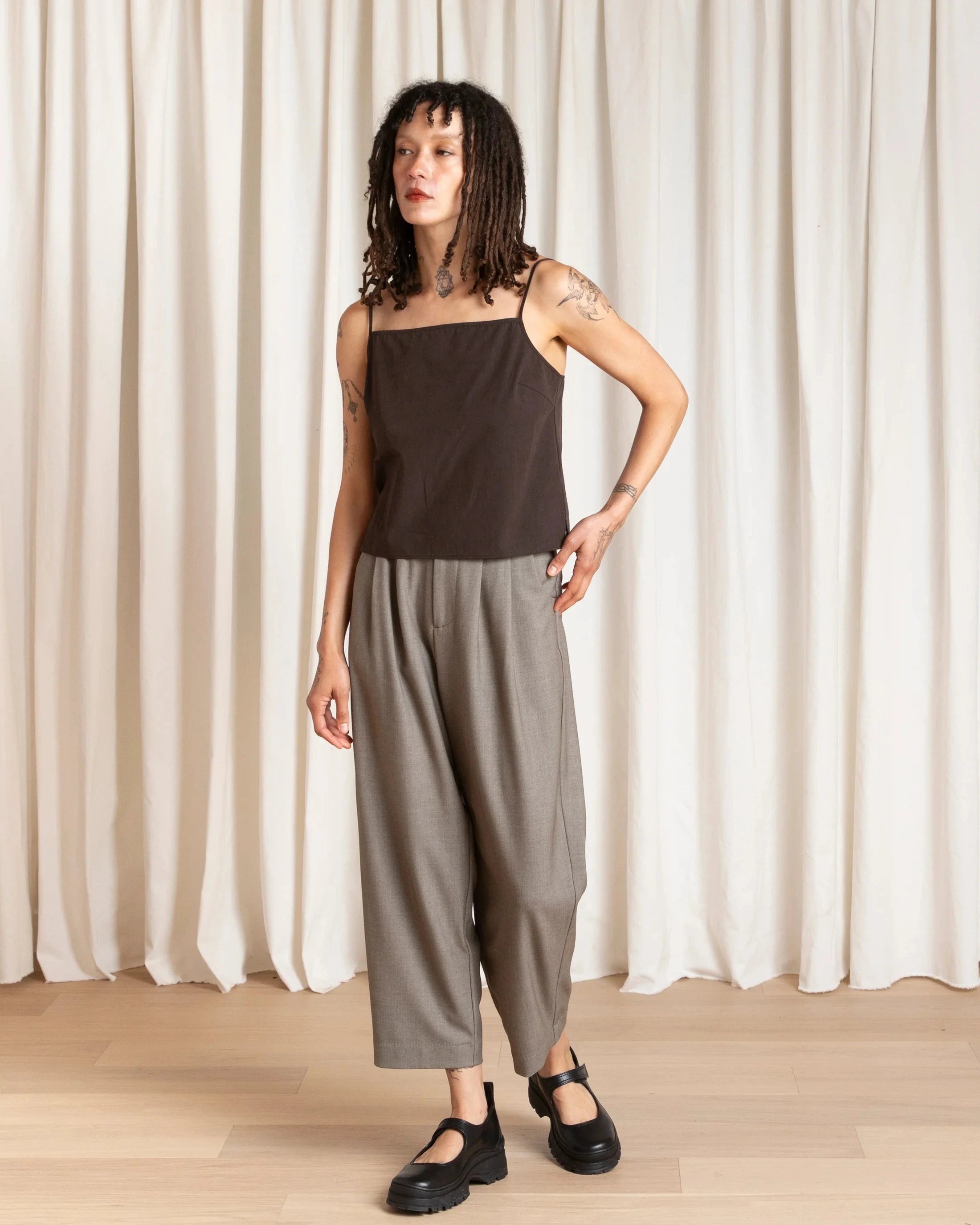 Woman wearing a brown top and gray pants standing against a white curtain.