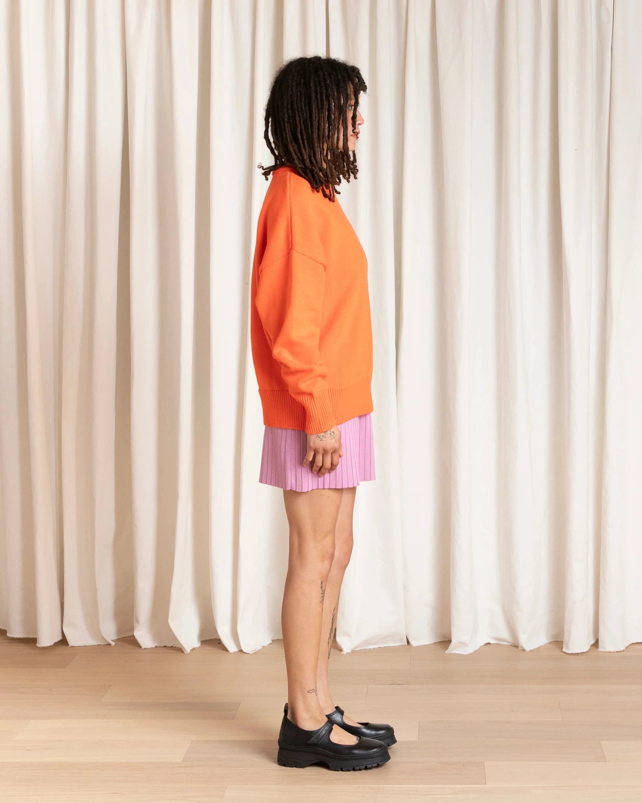 Person wearing an orange sweater and pink skirt standing in front of a white curtain.