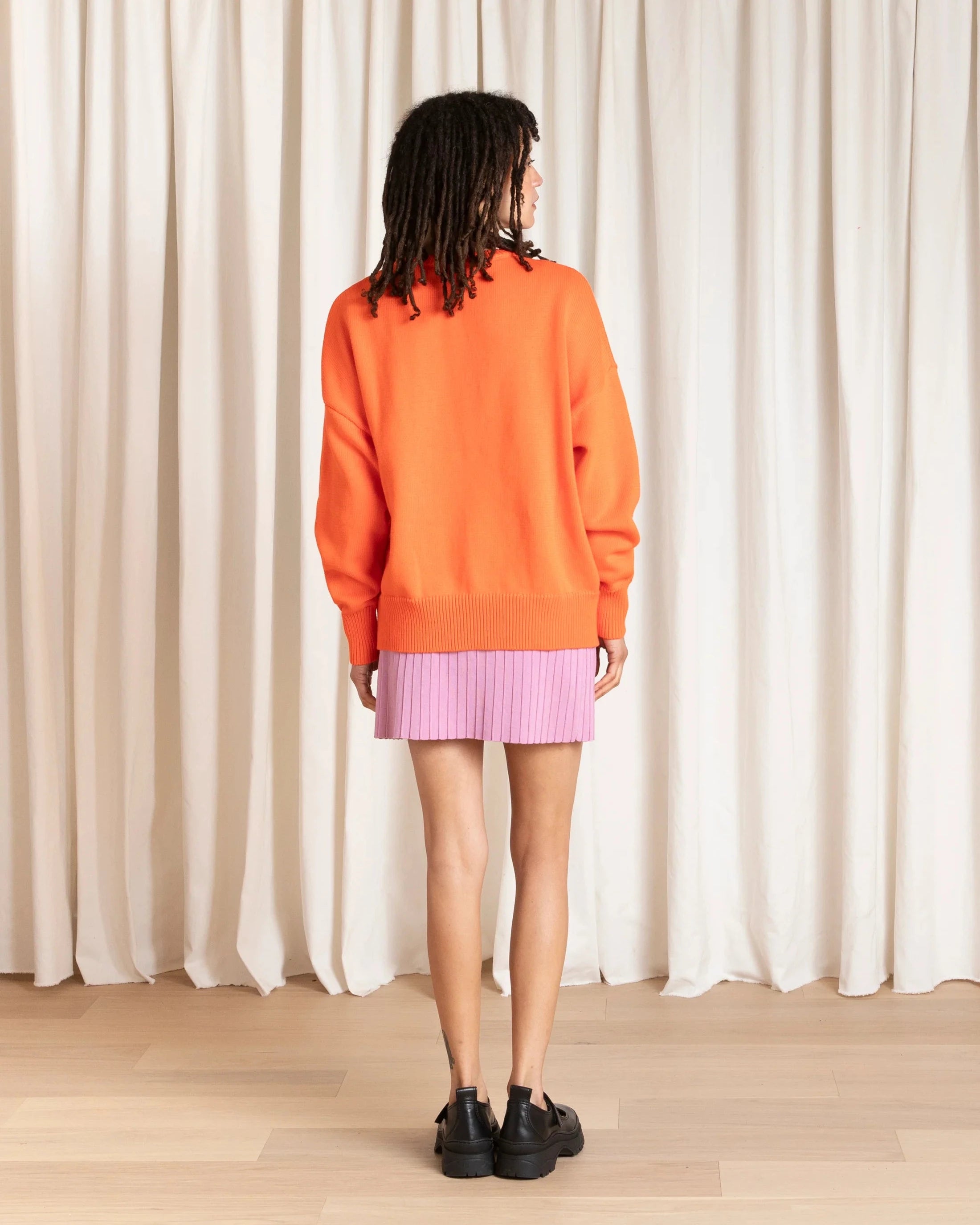 Person wearing an orange sweater and pink skirt standing in front of a white curtain.