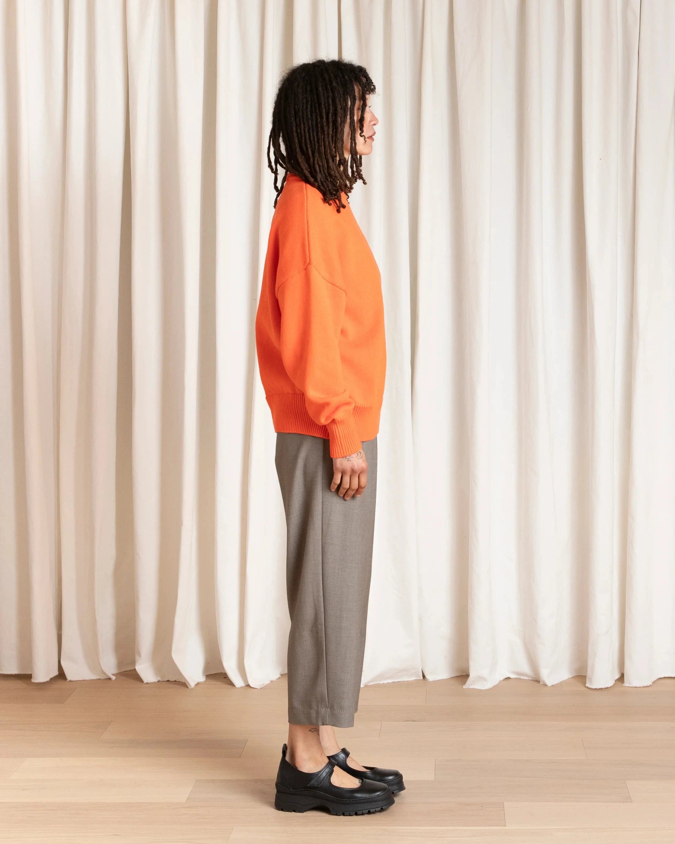 Person wearing an orange sweater and gray pants standing against a white curtain.