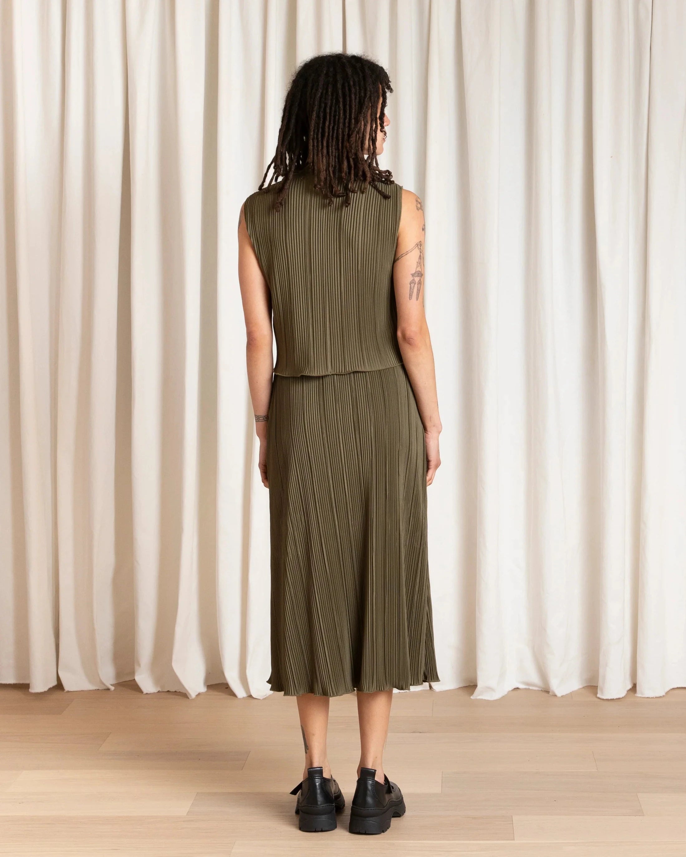 Person wearing a green sleeveless top and pleated skirt standing against a white curtain.