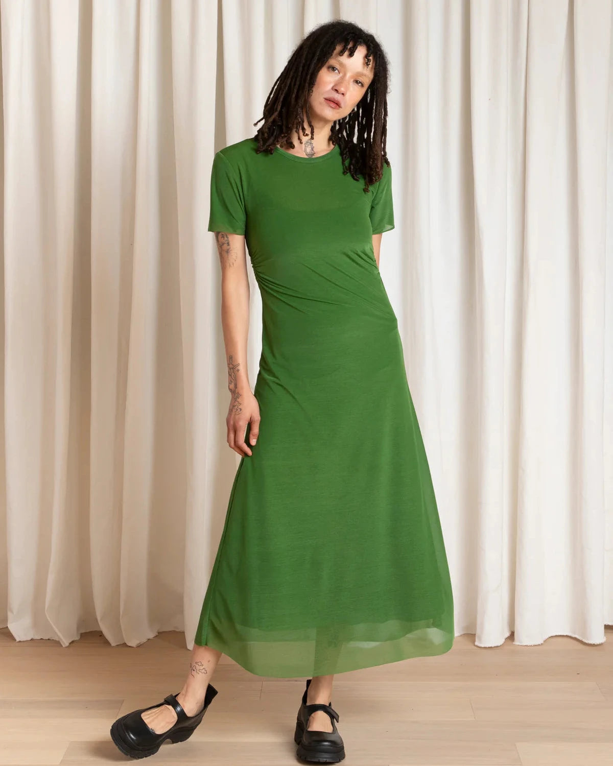 Woman wearing a green dress standing against a white curtain.