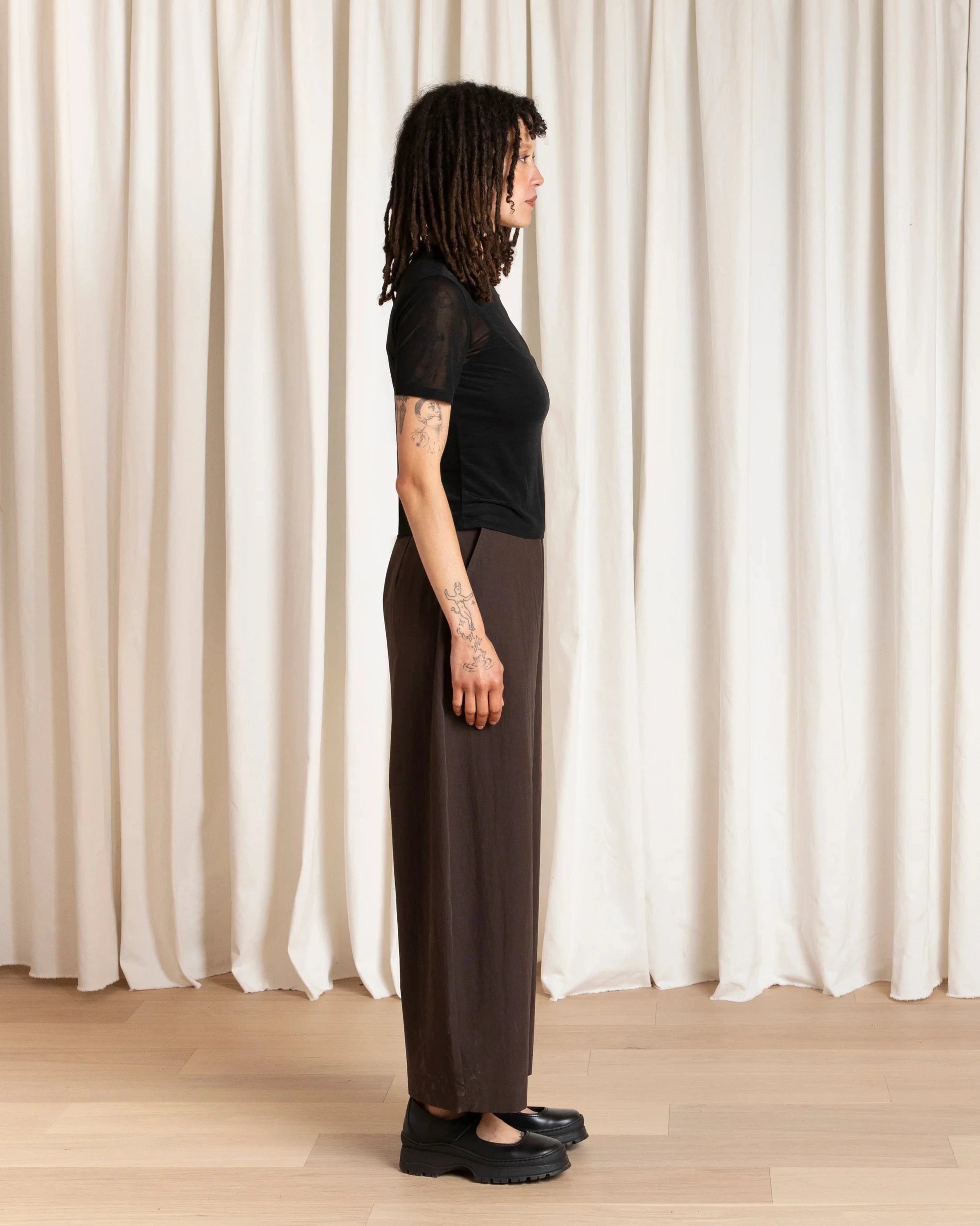 Person wearing a black top and brown pants standing against a white curtain.