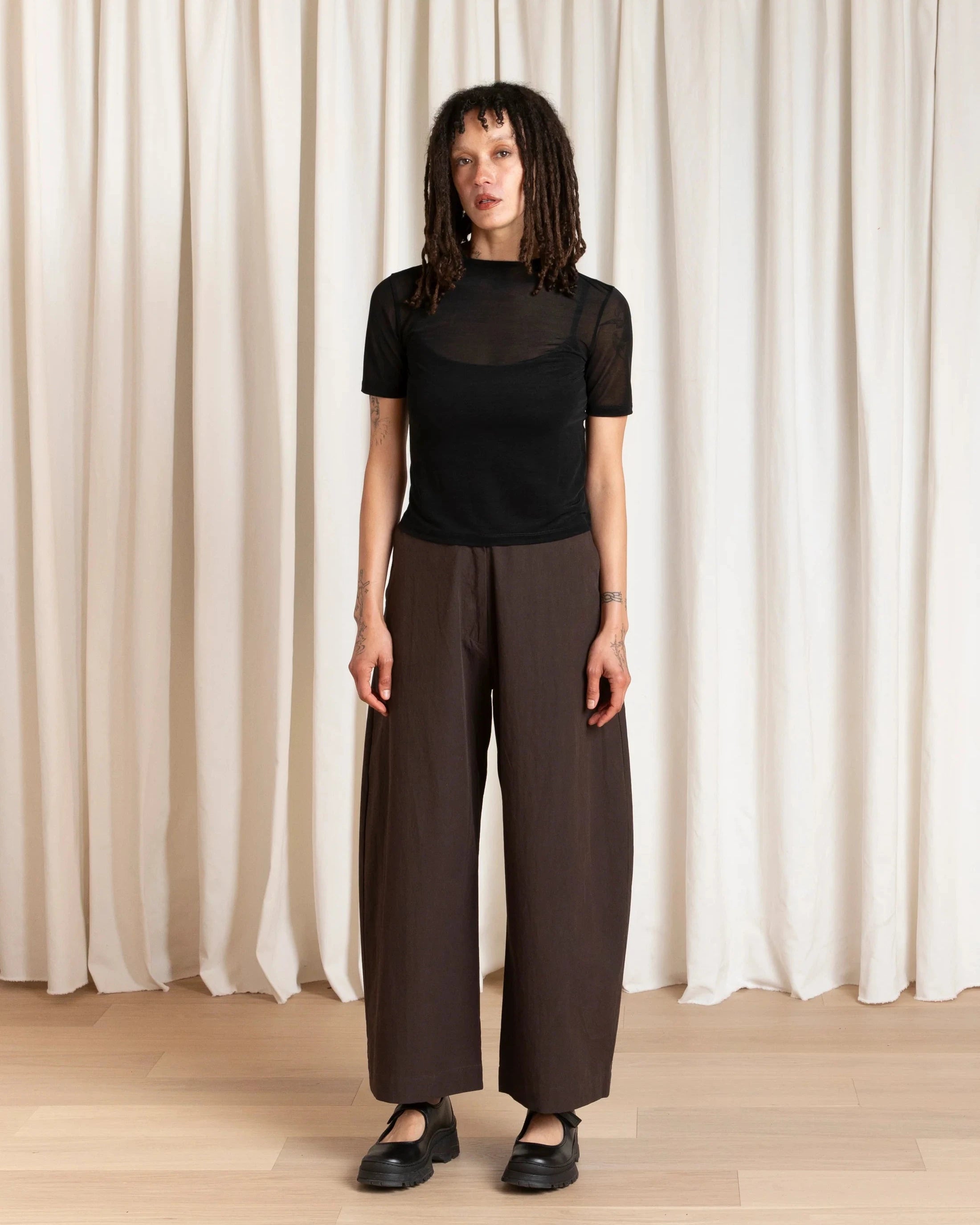 Person wearing a black top and brown pants standing against a white curtain.