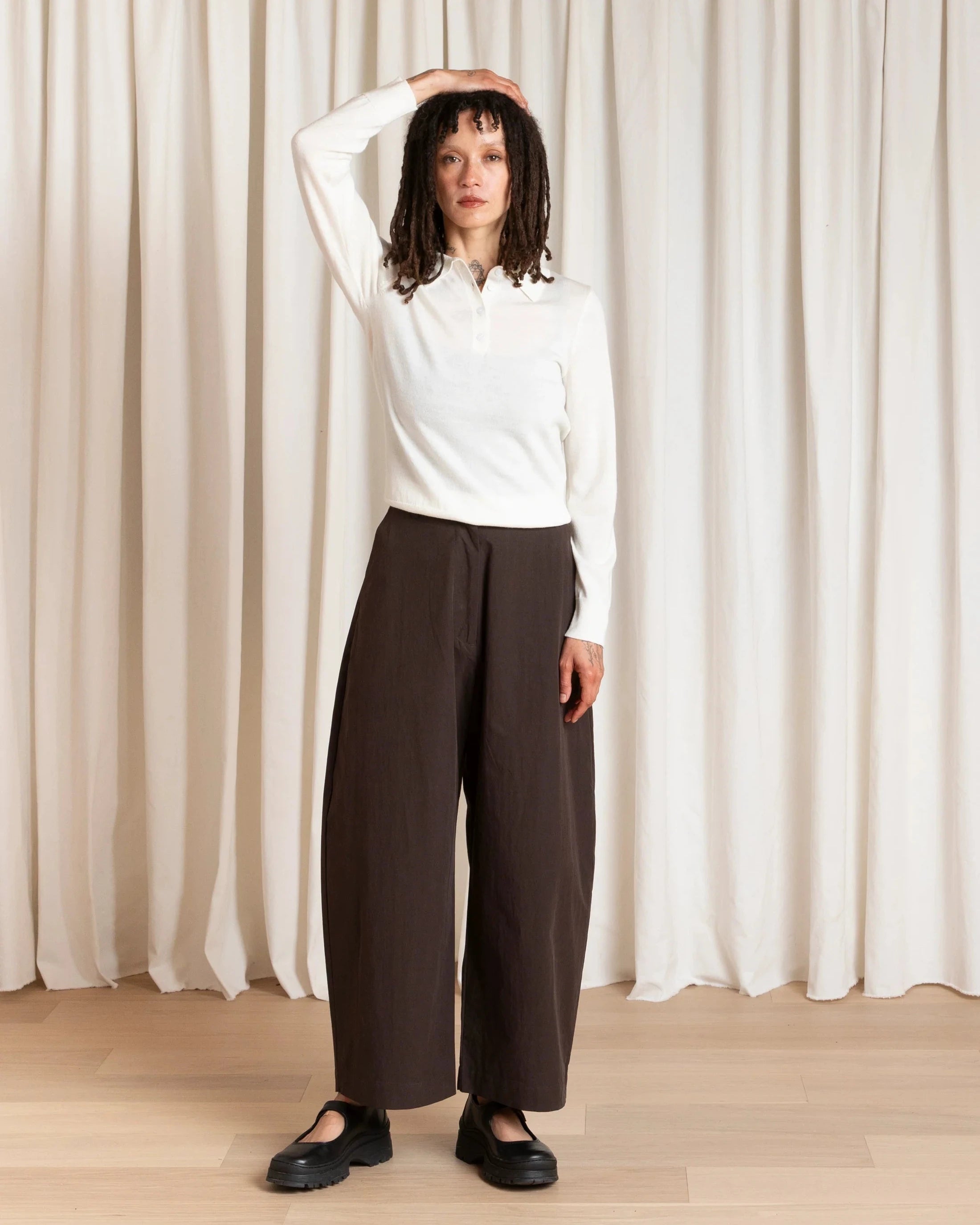Person wearing a white sweater and brown pants standing against a white curtain.