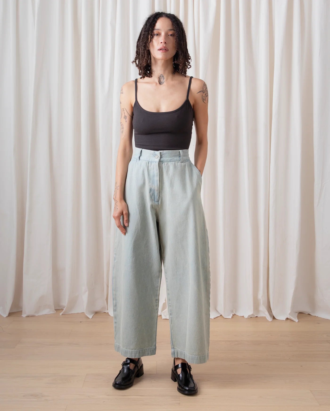Woman wearing a black tank top and light blue pants standing against a white curtain.