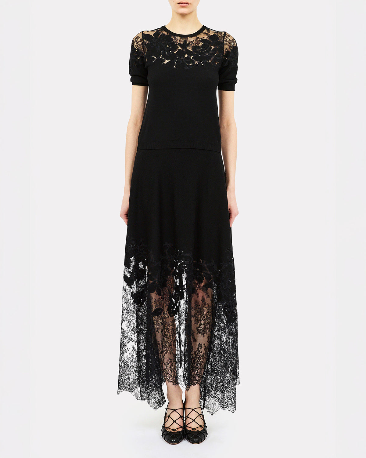 A model wearing a black short-sleeve maxi dress with sheer floral lace panels at the shoulders and a matching lace overlay on the skirt, paired with black lace-up heels against a plain white background.