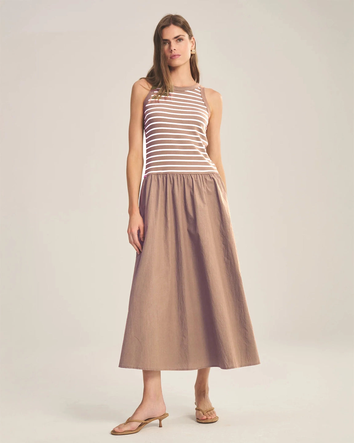 Woman wearing a dress that has a striped sleeveless top and a long beige skirt on a plain background