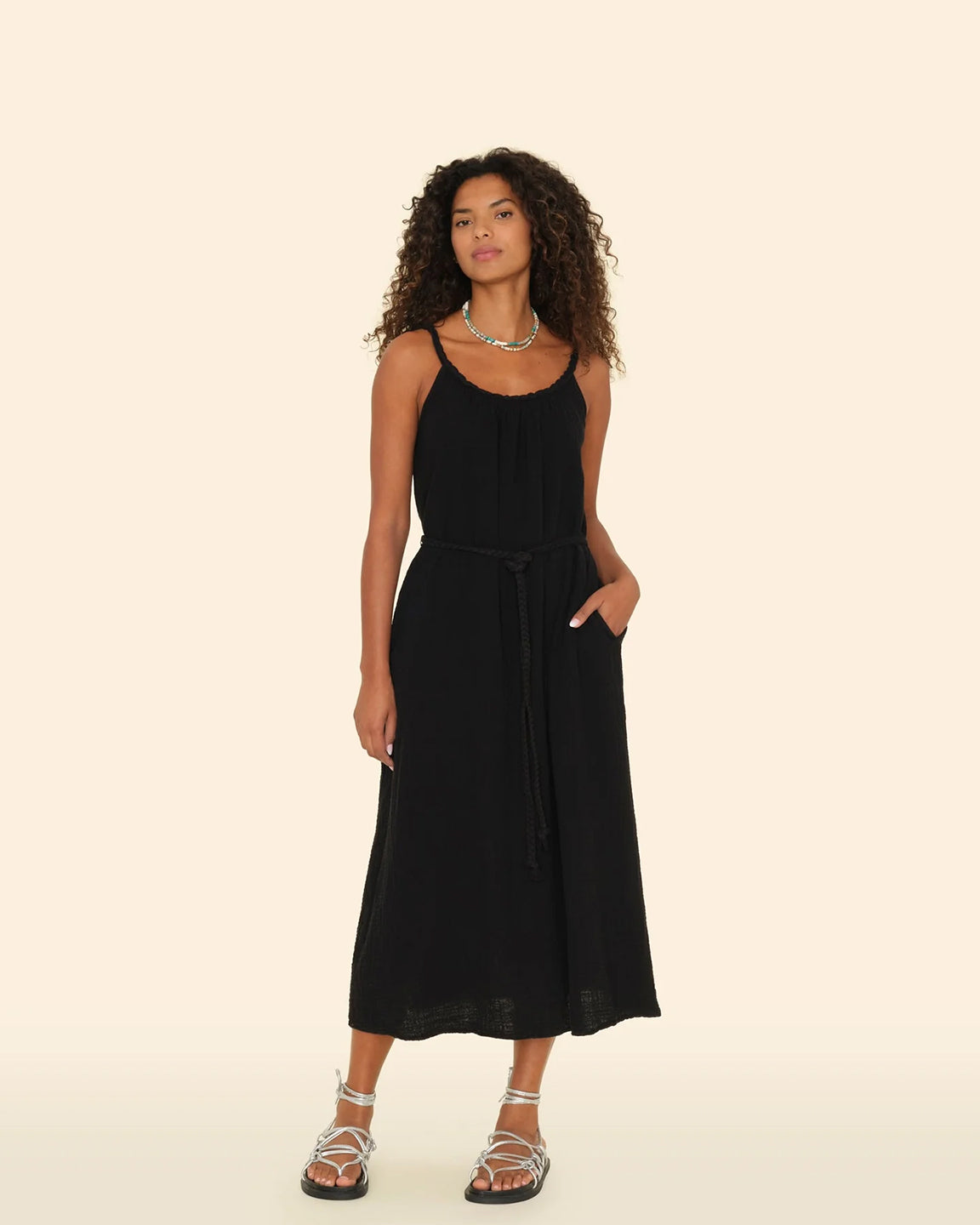 A woman standing in a black midi dress with a twisted braid neckline and straps, side seam pockets, and a braided self-tie belt.