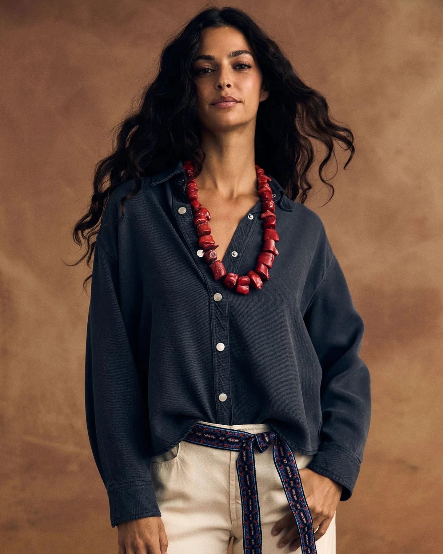 Woman wearing a dark shirt and beige pants with a red necklace against a brown background