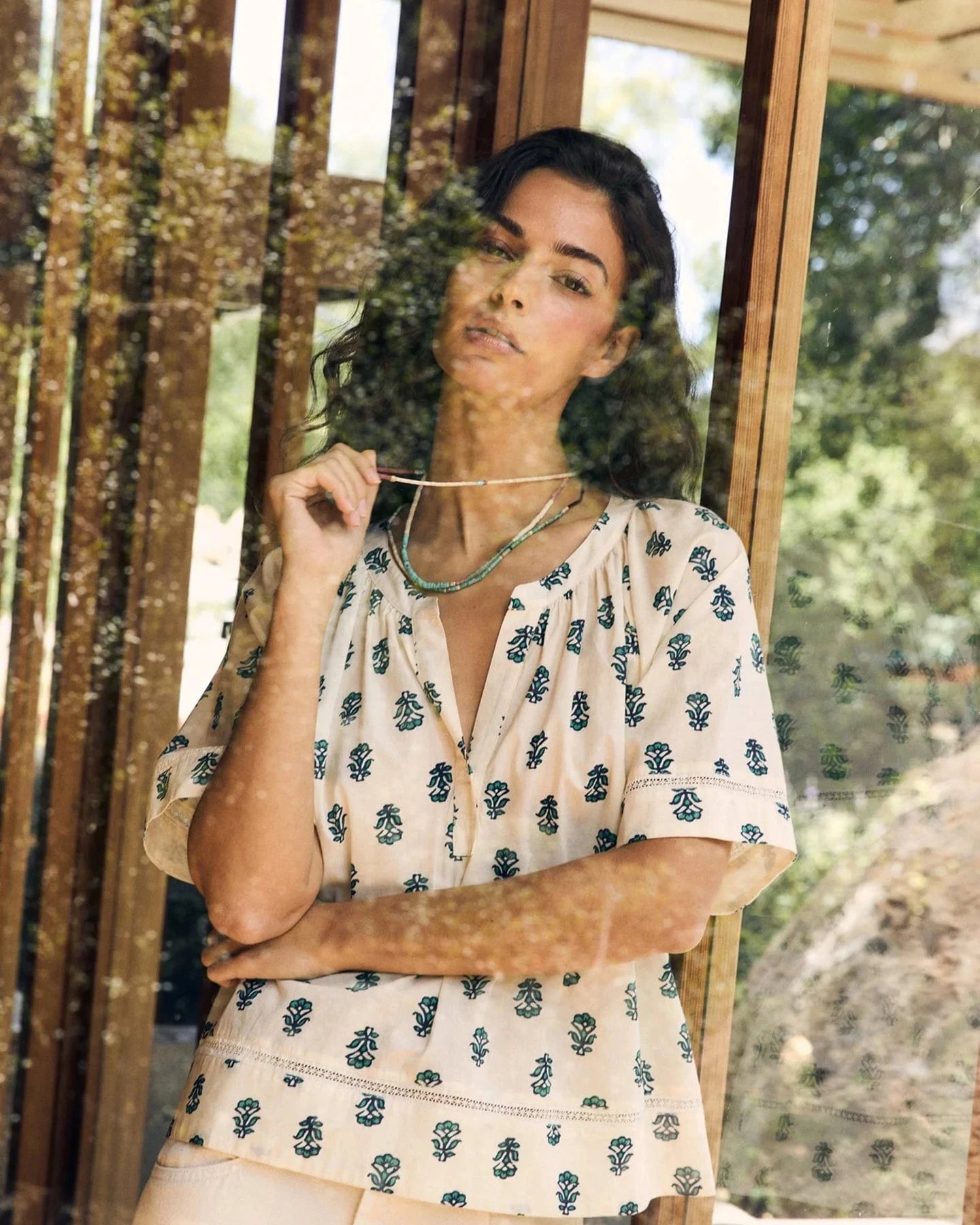 Woman wearing a patterned blouse standing in front of a glass door with greenery outside.