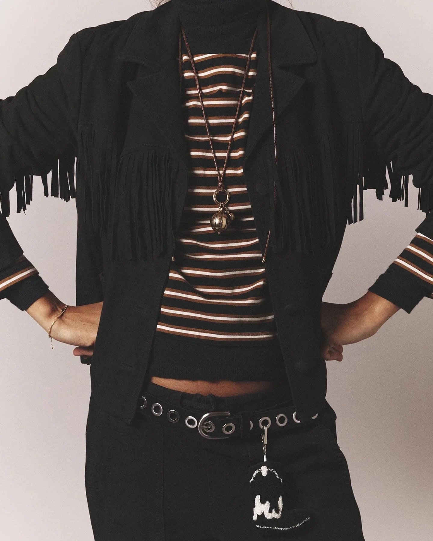 Person wearing a black jacket with fringe, striped top, and black pants on a plain background