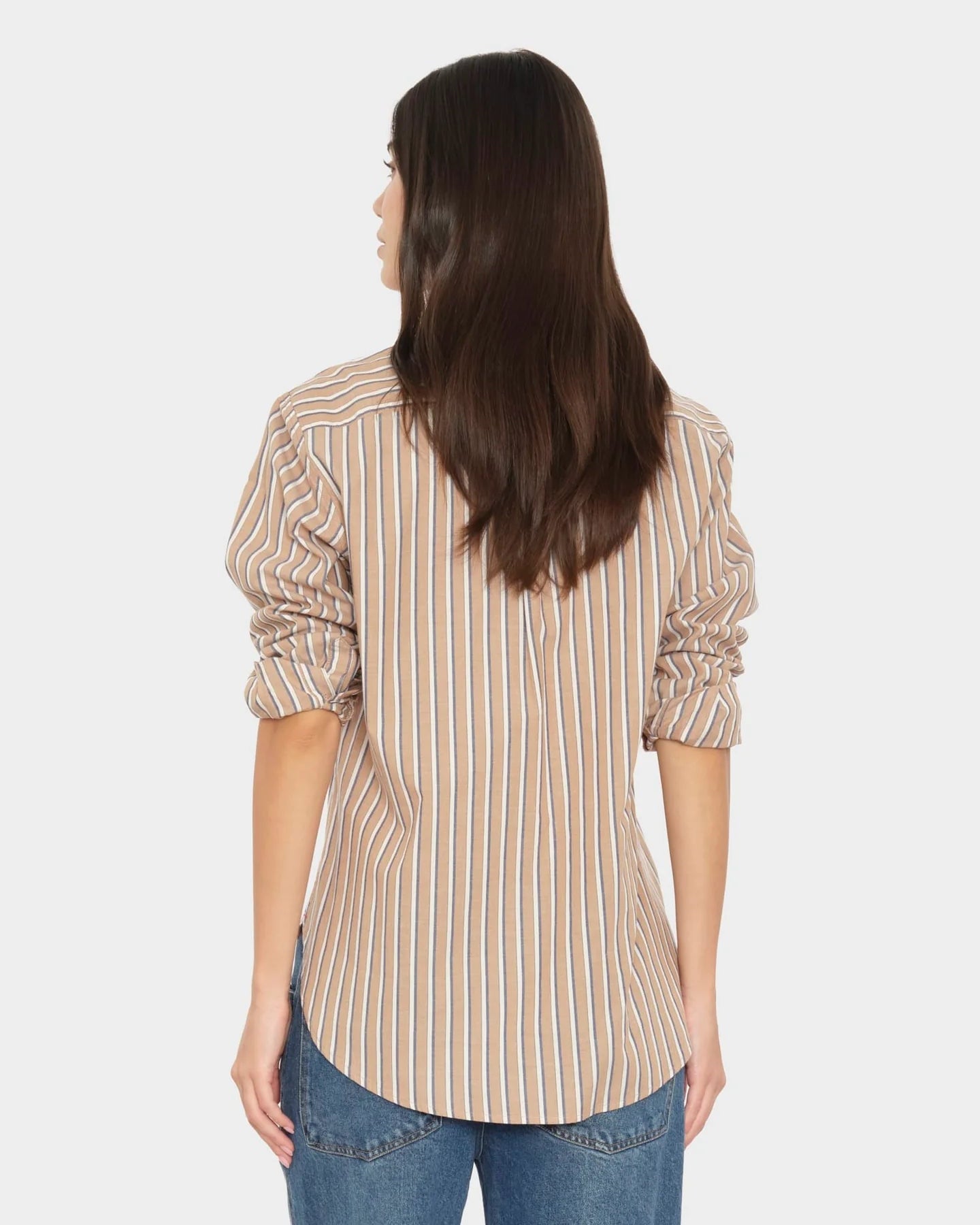 A woman viewed from the back wearing a beige and white vertically striped long-sleeve blouse with gathered cuffs, paired with blue jeans, against a light gray studio background.