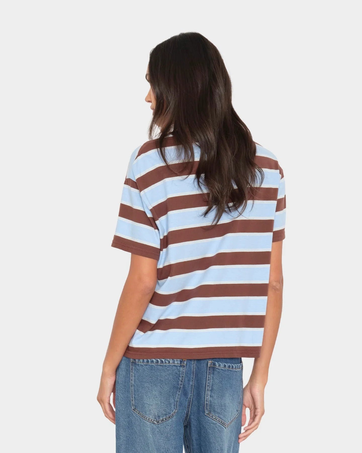 A woman shown from the back wearing a brown and light blue horizontally striped short-sleeve t-shirt and blue jeans, against a light gray studio background.
